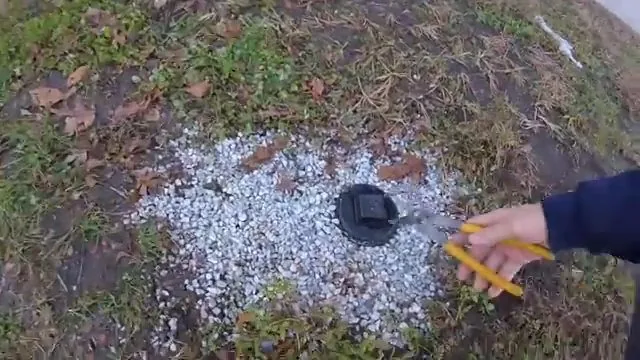 Black cleanout cap in a bed of white gravel, with a hand pointing at it using a yellow pencil.