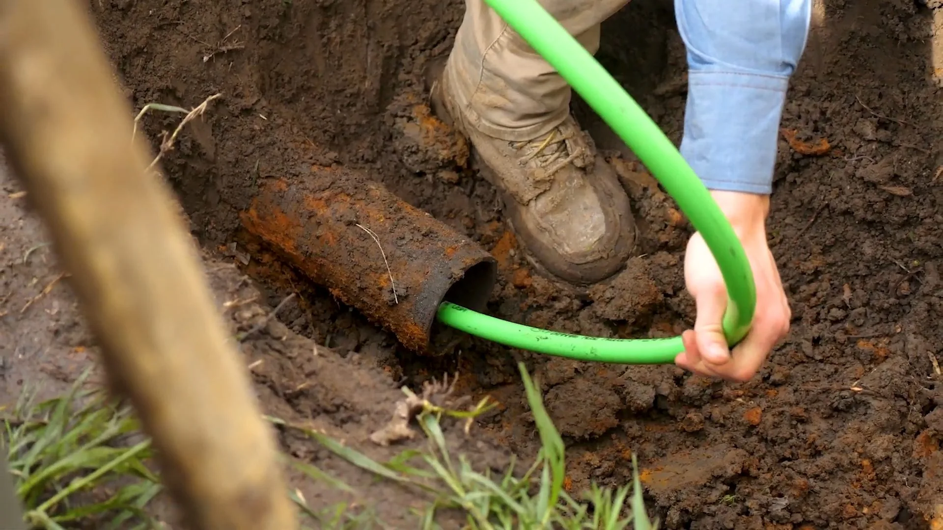 Person in boots pulling a green flexible line out of the end of a rusted pipe in a dirt trench.