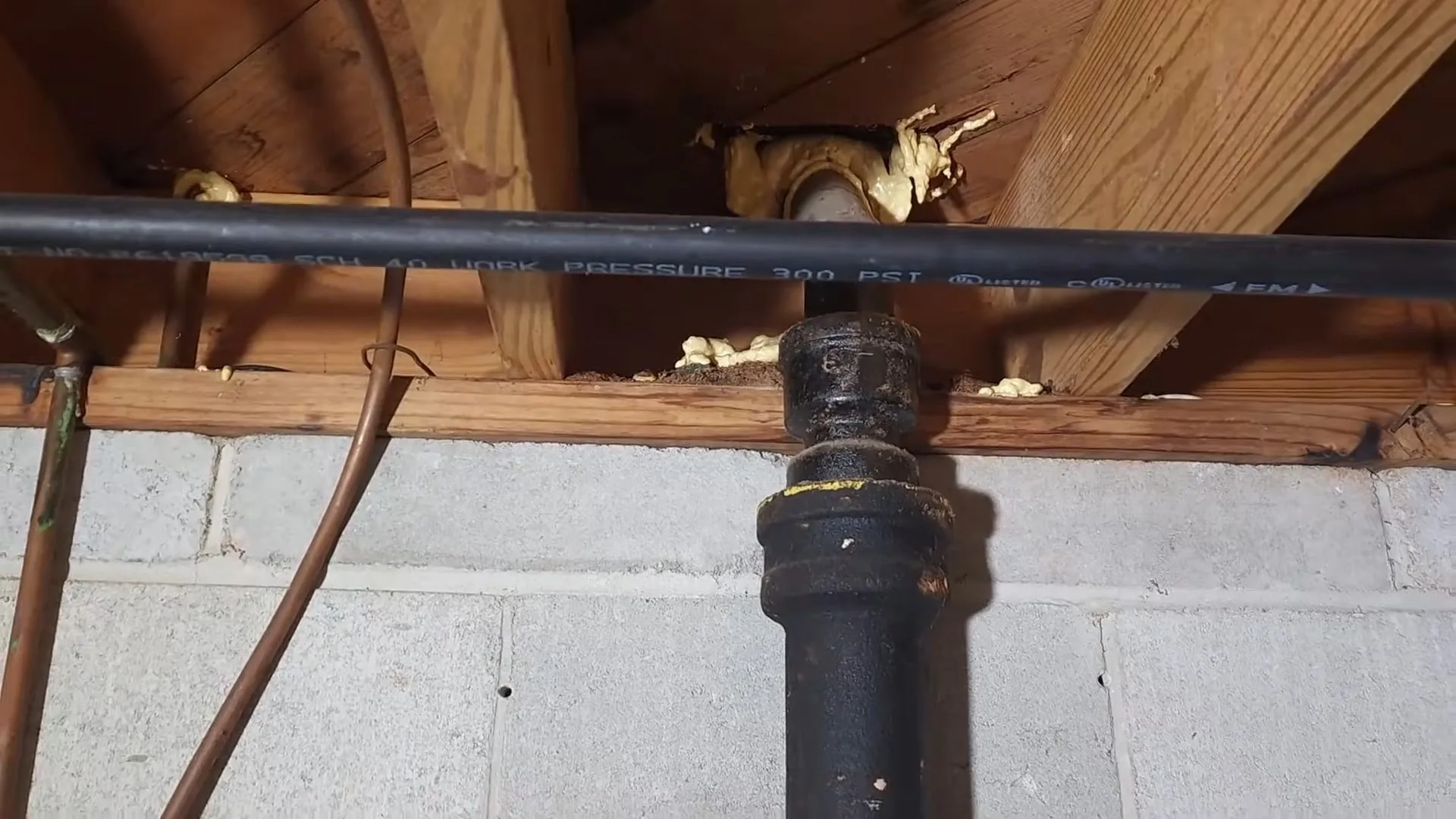 Basement joists above a concrete block wall with a black cast-iron drain stack, a black pressure-rated water line and a copper pipe.