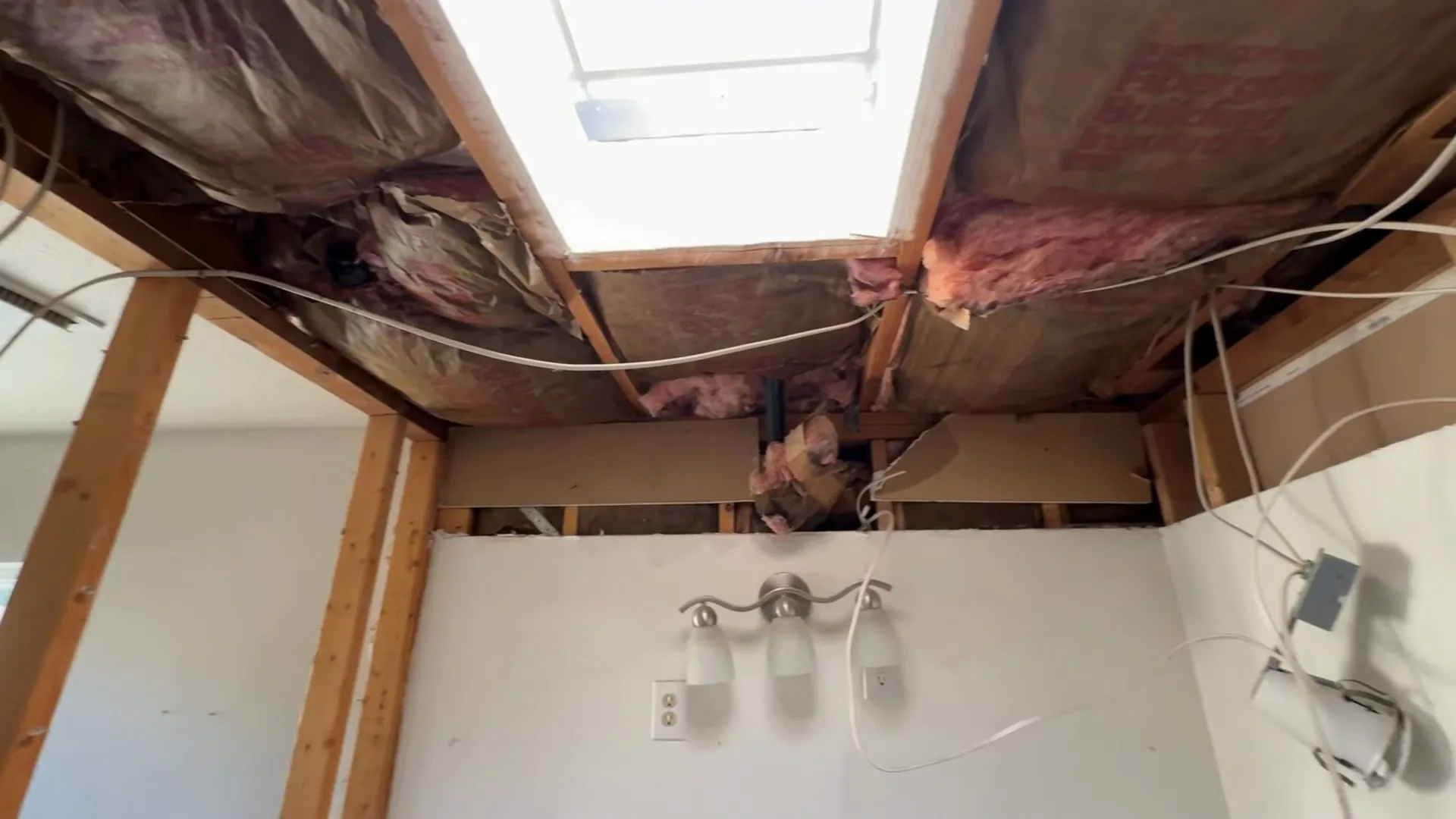 Ceiling opened to the rafters and pink insulation above a wall with a three-bulb vanity light.