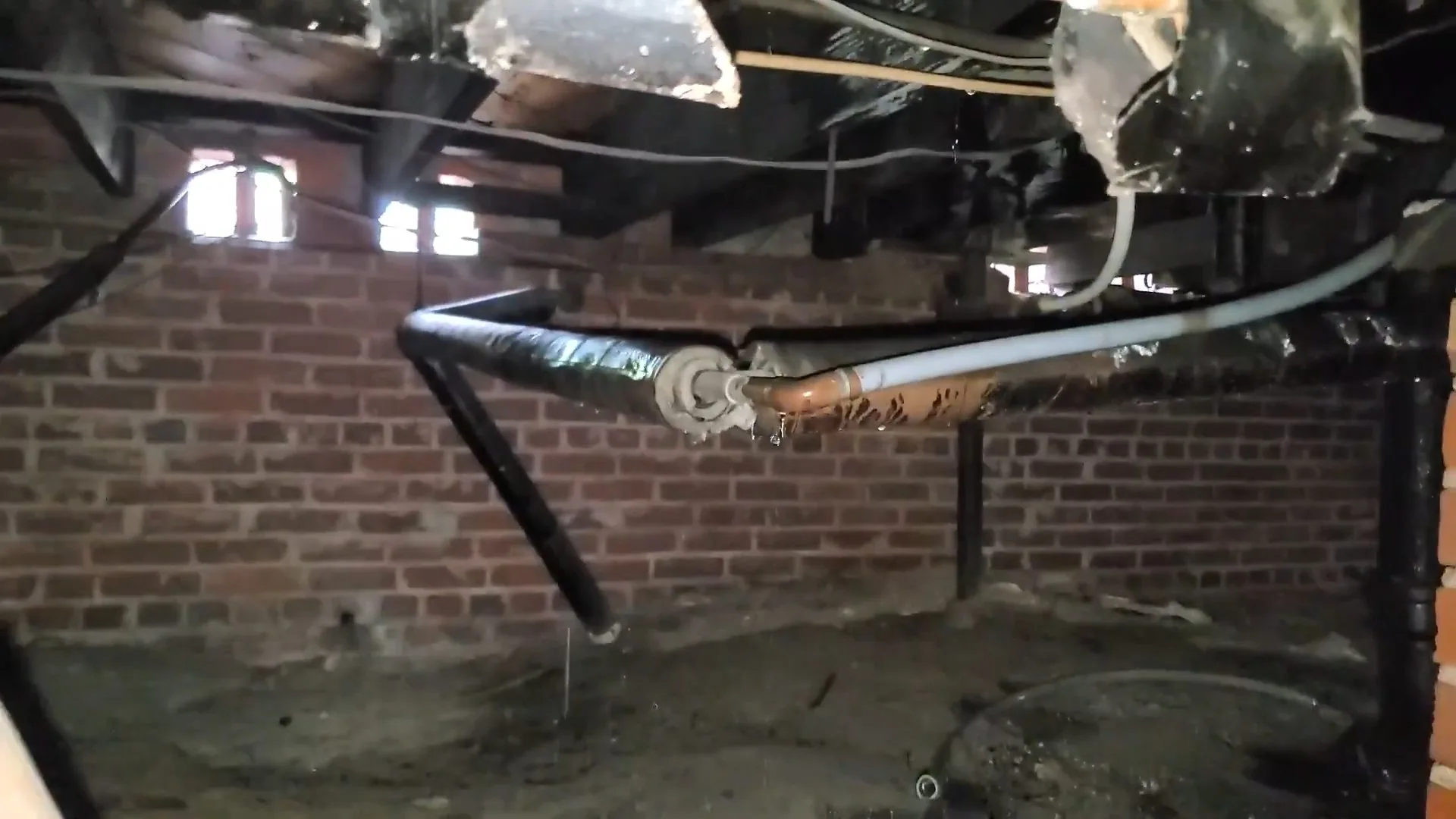 Crawl space with a brick foundation wall and black and white pipes angling down from the floor above.