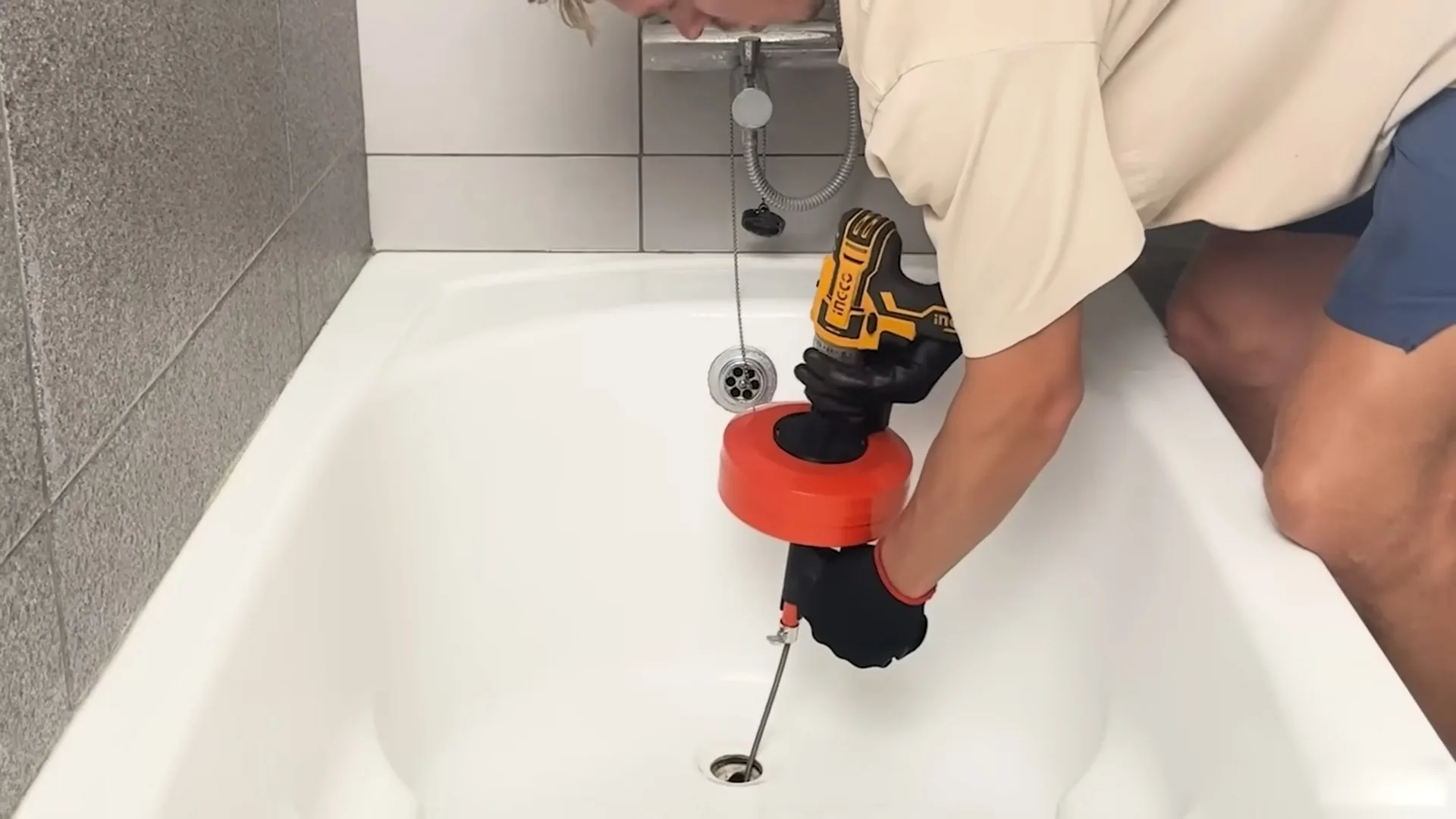 Person using a drill-powered drain snake with a red drum at a white bathtub drain.