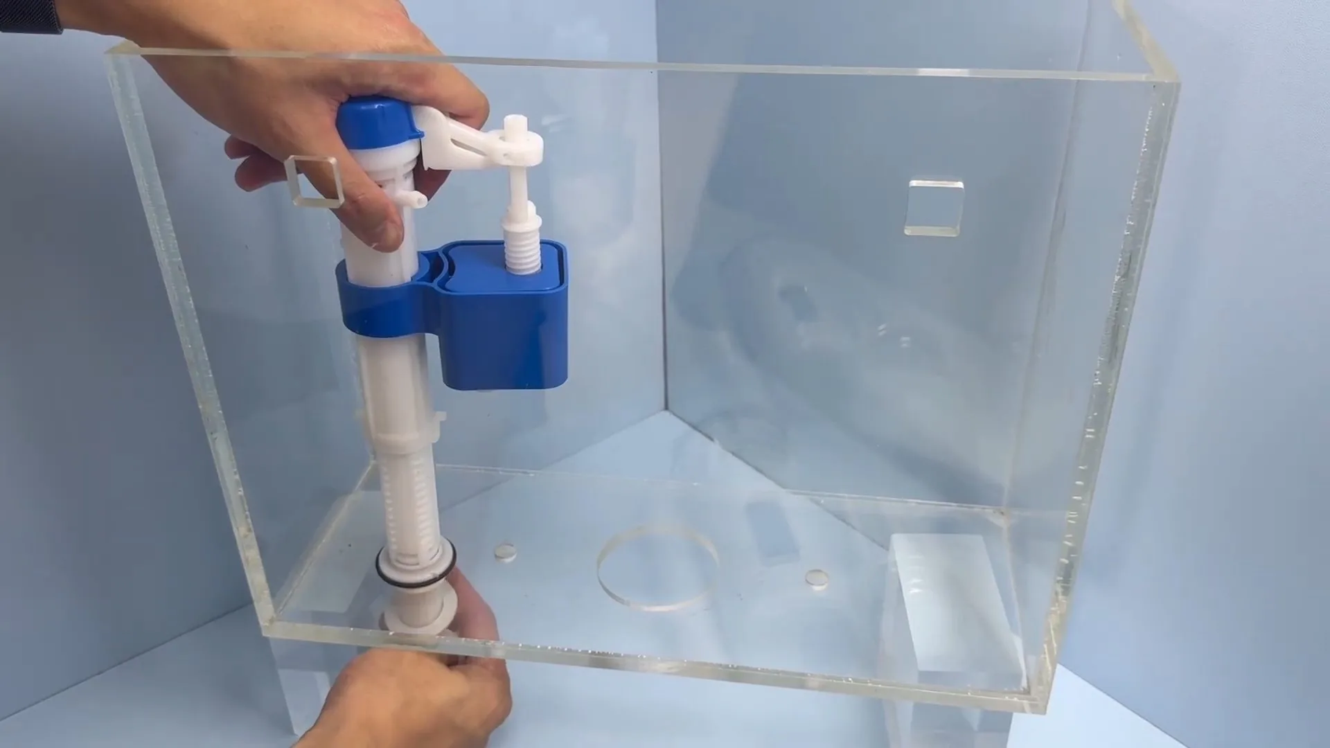 Clear acrylic toilet tank showing a fill valve with a float, with a hand holding its base.