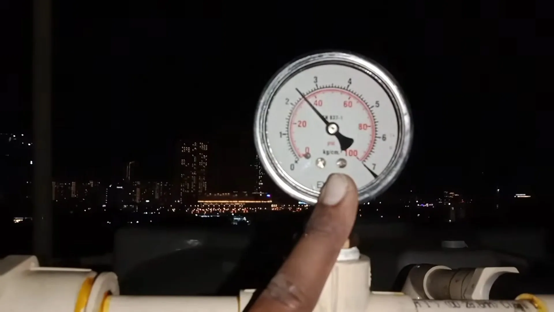 Finger pointing at a pressure gauge on a white pipe at night, with city lights in the background.