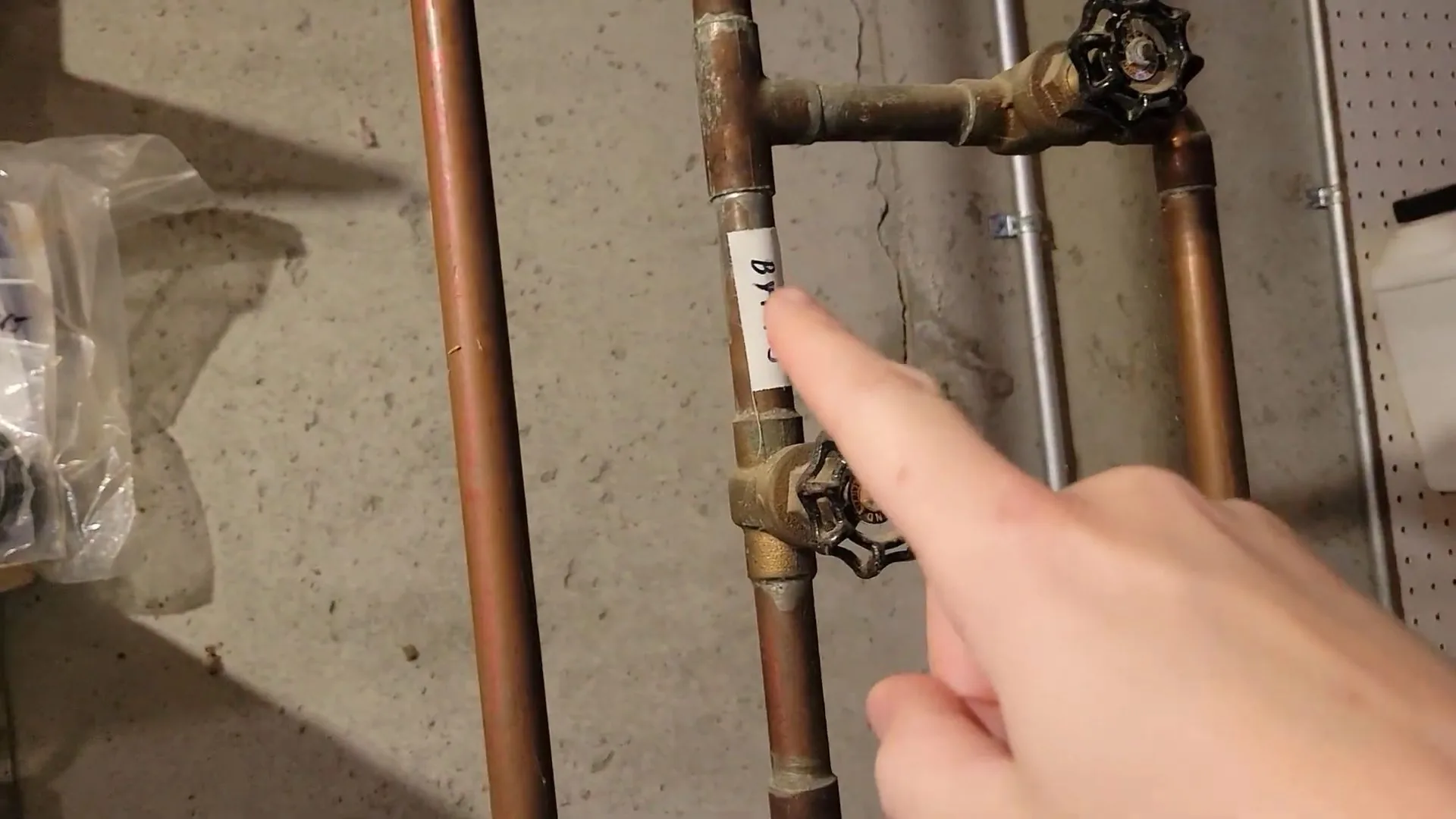 Finger pointing at a shutoff valve on copper pipes beside a basement wall.