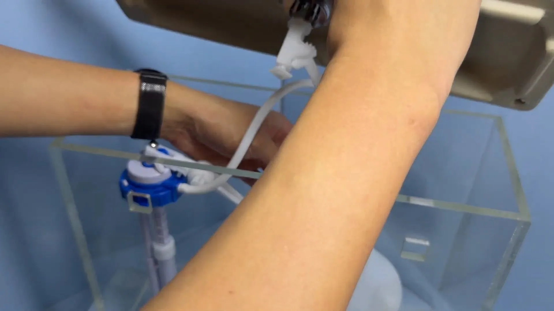 Forearm reaching into a clear acrylic toilet tank to adjust a blue fill valve.