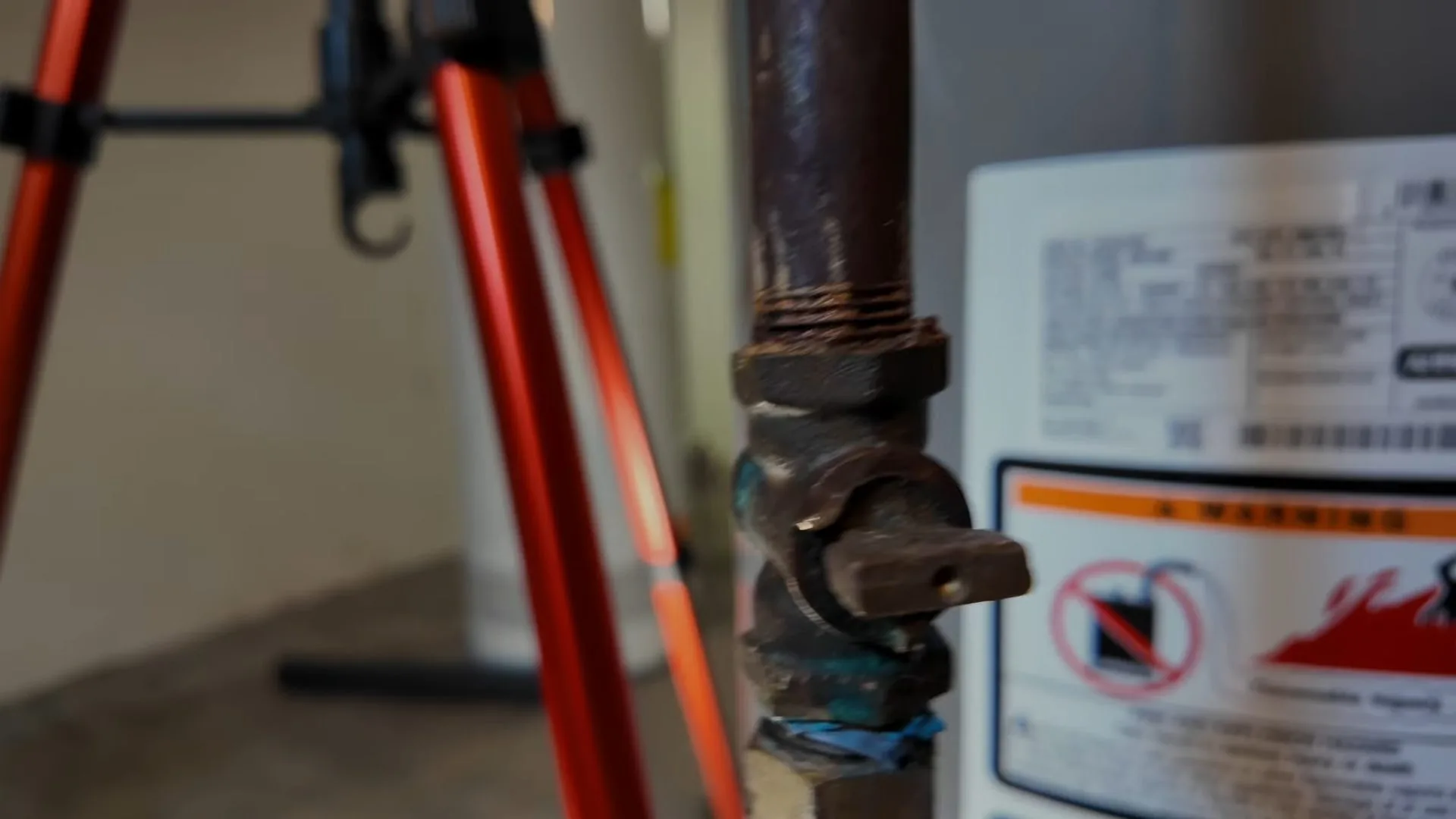 Gas shutoff valve on a pipe beside a water heater with a warning label, and an orange tripod behind it.