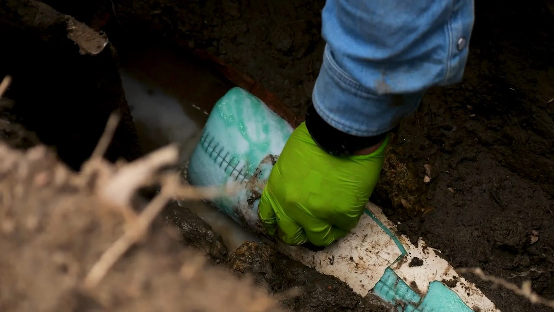 Green-gloved hand pressing a teal flexible liner against a pipe in a dirt trench.