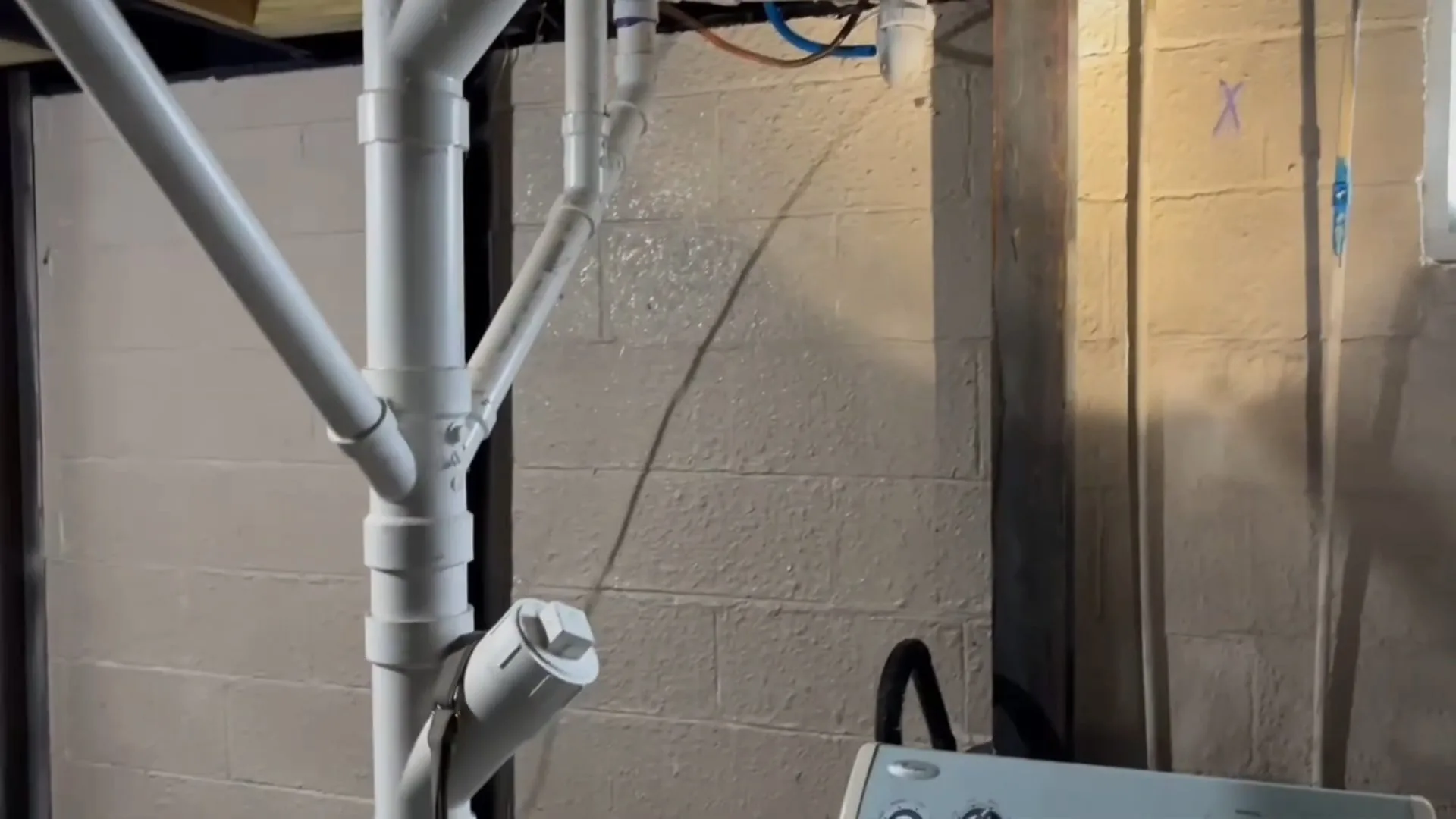 Gray PVC drain stack with branch fittings against a concrete block wall in a basement.
