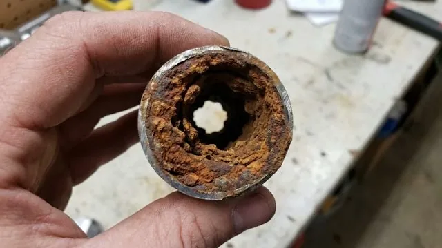 Hand holding a short section of old pipe packed with orange rust on the inside.
