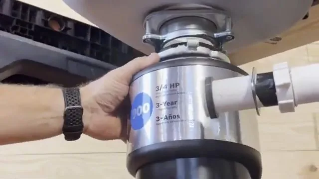 Hand holding a stainless steel garbage disposal mounted under a sink with a drain pipe attached.