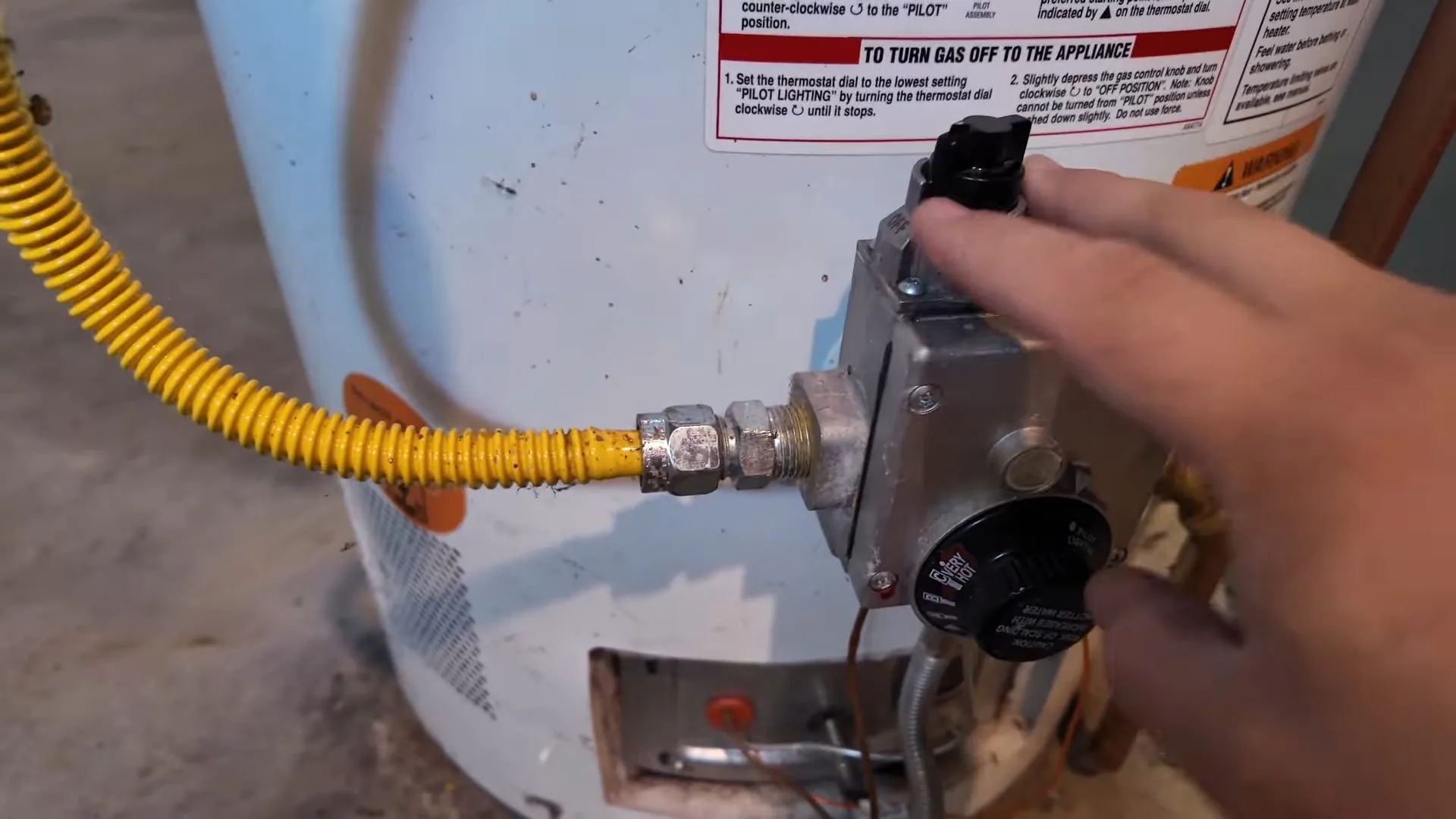 Hand reaching to the gas control on a white water heater, with a yellow flexible gas line attached.