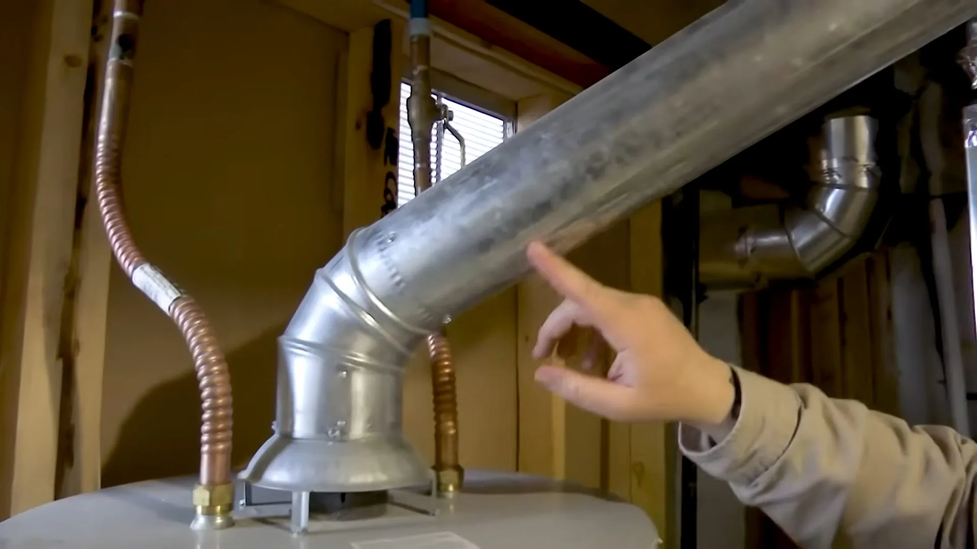 Hand pointing along a metal vent pipe that rises from a water heater in a utility room.