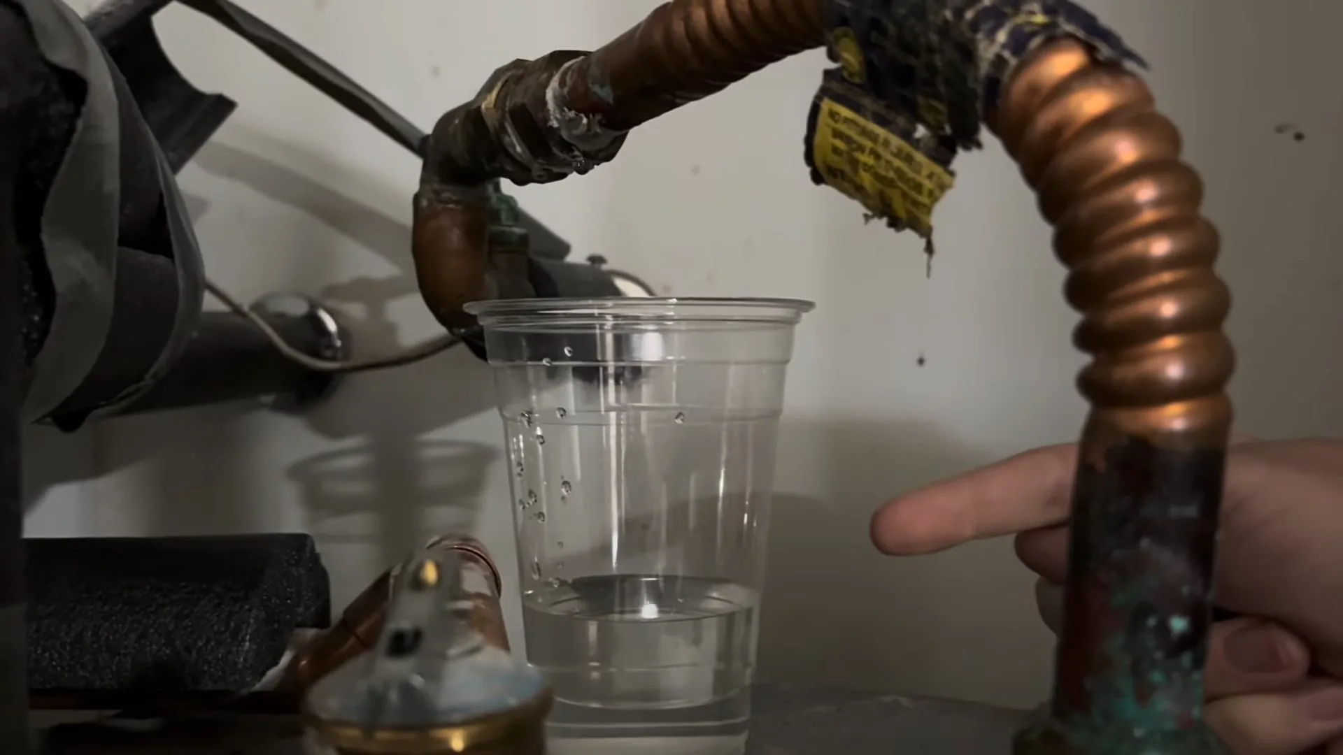 Hand pointing at a corroded copper and corrugated connector dripping into a clear cup.