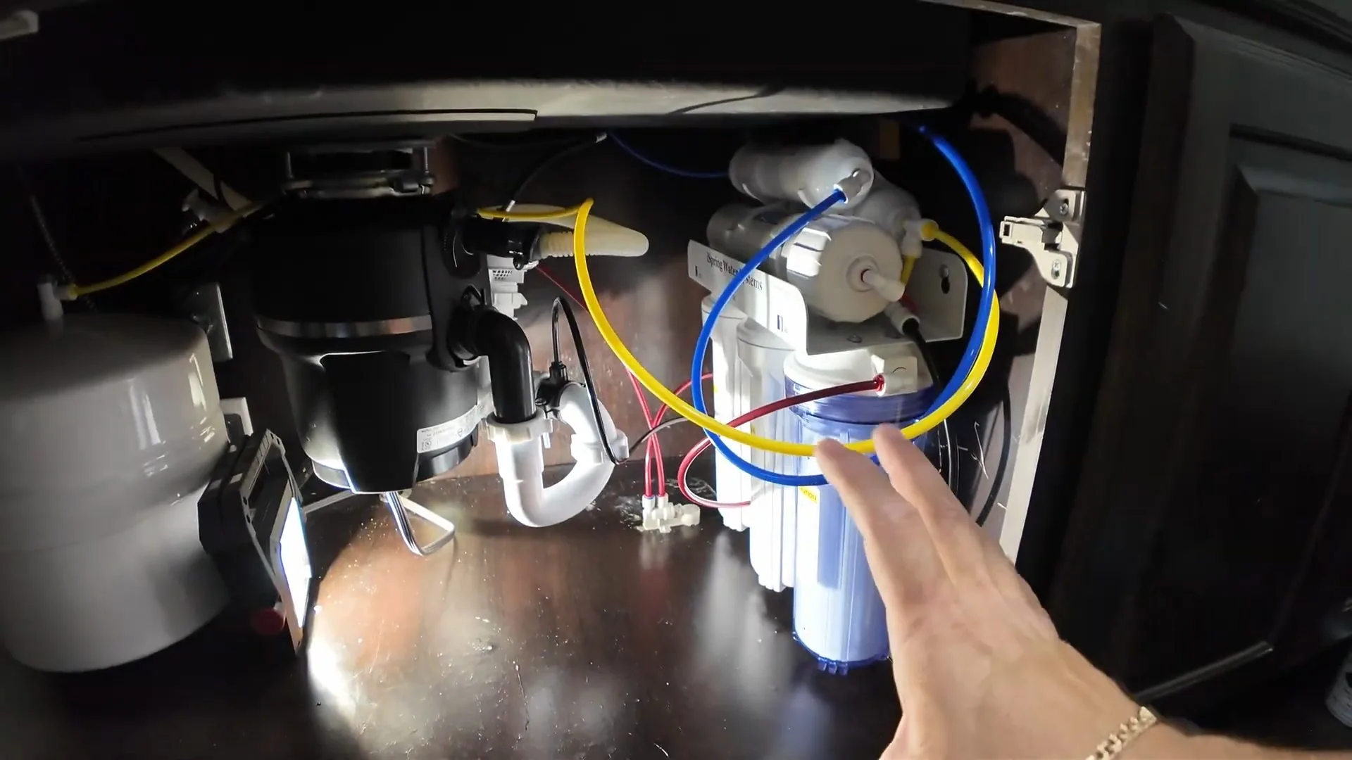 Hand pointing at a set of white reverse osmosis filters with yellow and blue tubing under a kitchen sink, beside a garbage disposal.