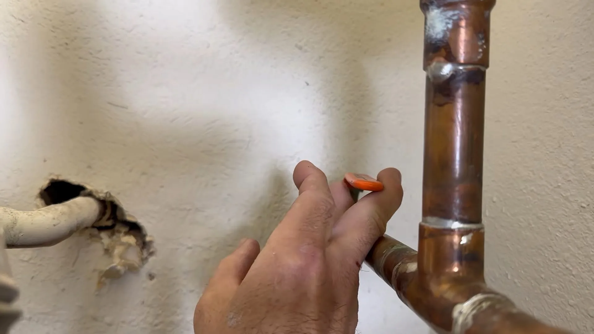 Hand holding an orange marker beside a copper pipe elbow, with a rough hole in the painted wall behind it.
