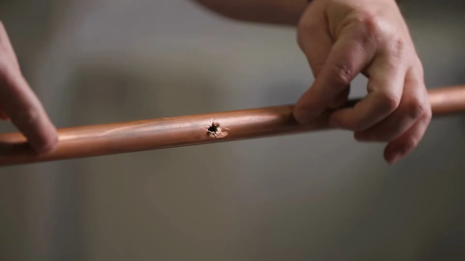 Two hands holding a length of copper pipe with a small pinhole in its side.