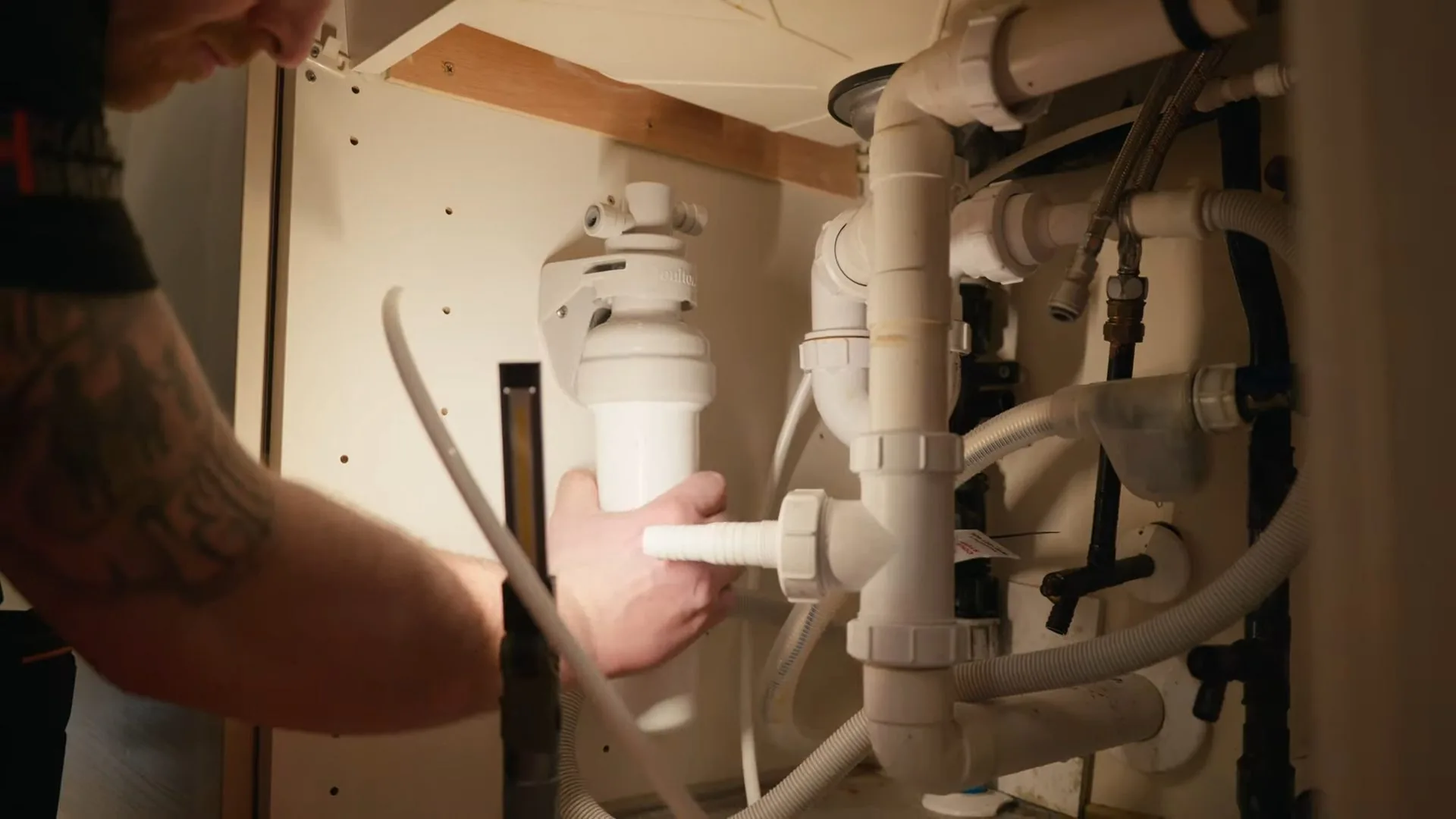 Man's hands holding a white filter housing under a kitchen sink, among drain pipes and tubing.