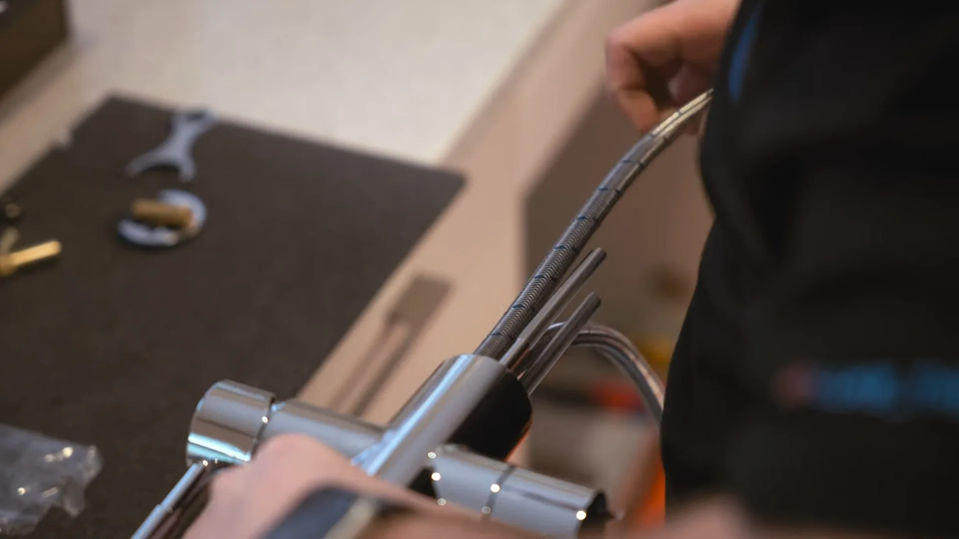 Hands installing a chrome faucet with a flexible supply line, with a wrench and small parts on a dark mat.
