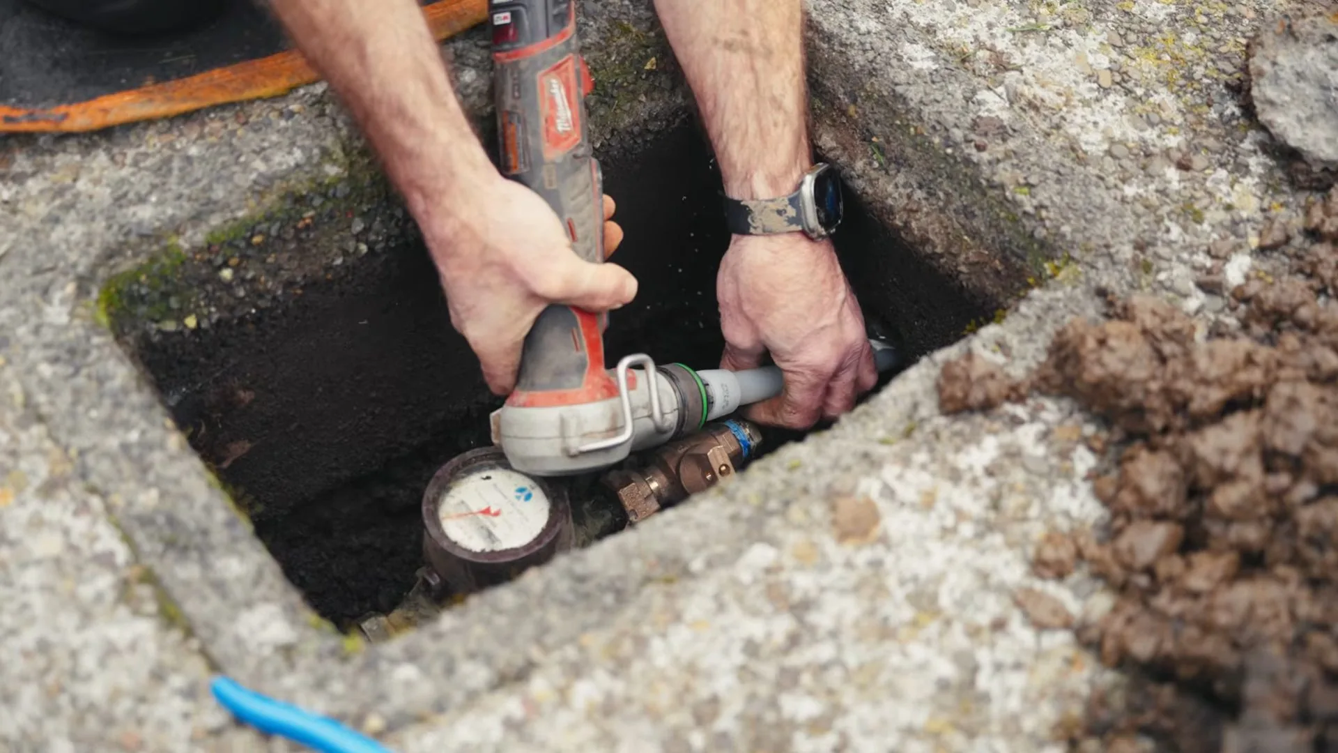 Two hands working on a pipe fitting next to a water meter inside an in-ground concrete meter box, with a red power tool held in one hand.