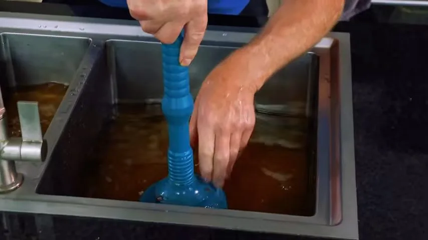 Man's hands working a blue plunger in a stainless steel sink filled with brown water.