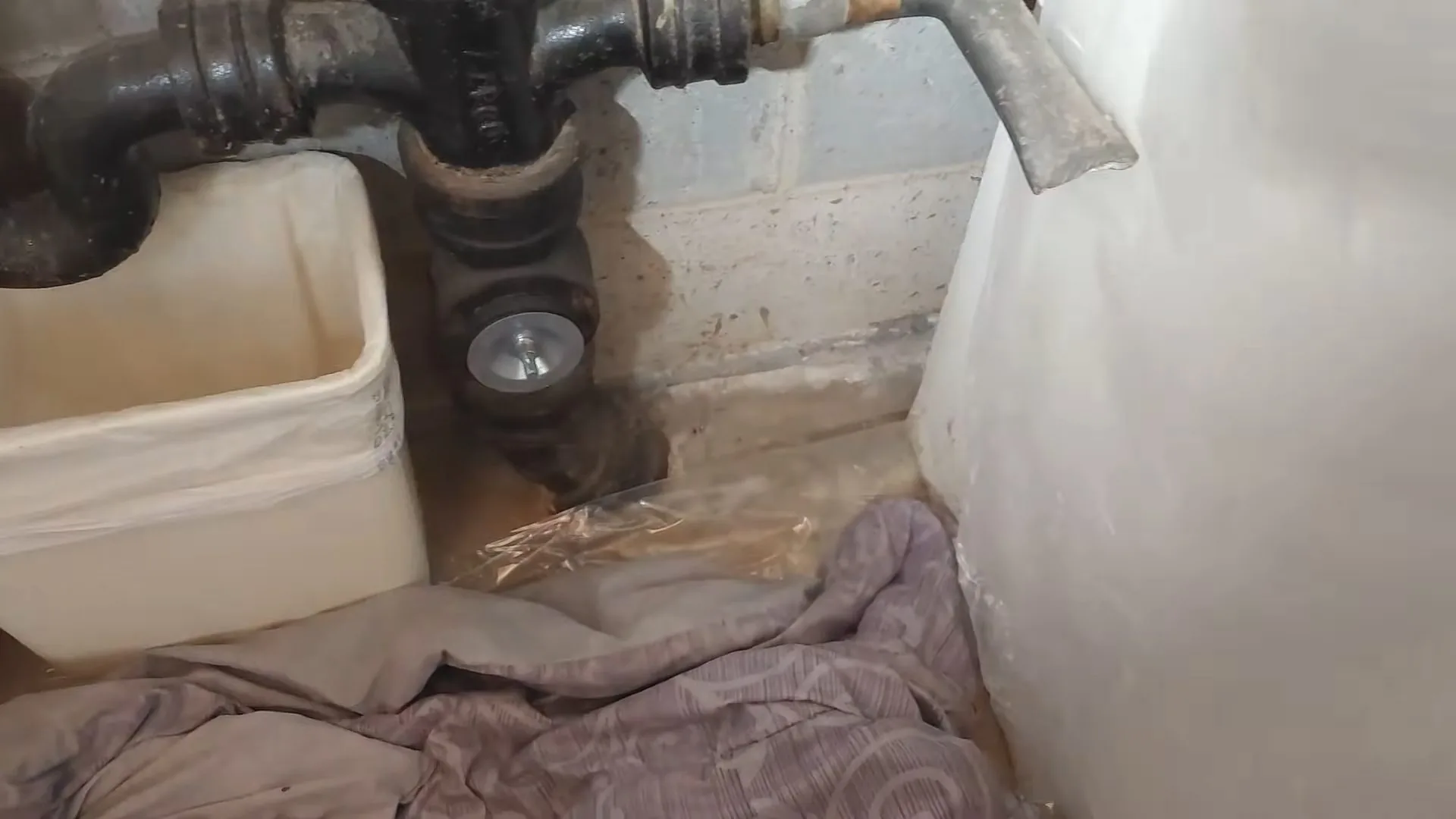 Dark iron drain piping with a round cleanout plug beside a white tub, with a cloth and a bin on the floor.