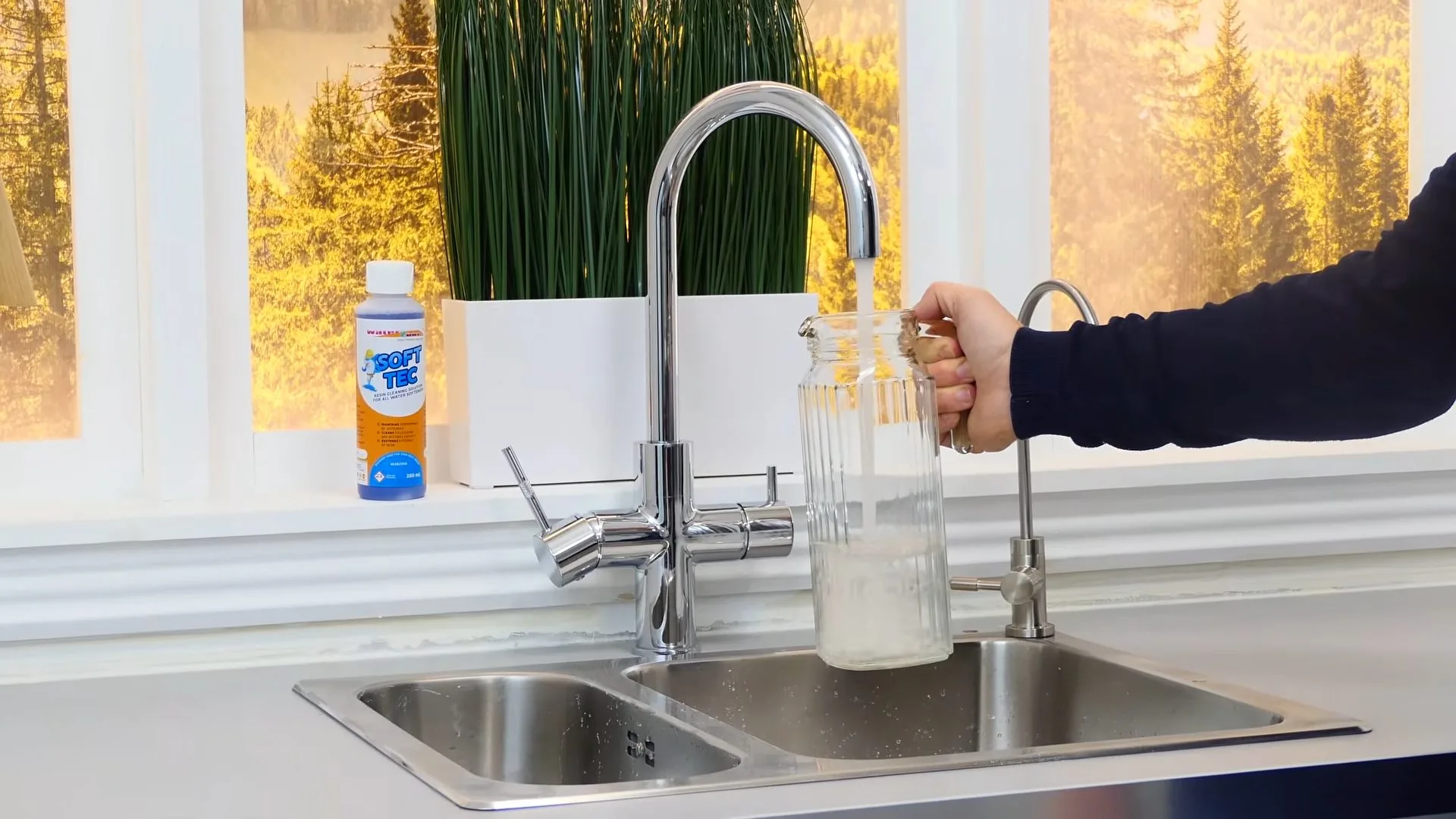 Hand holding a glass jar under a kitchen faucet beside a window.