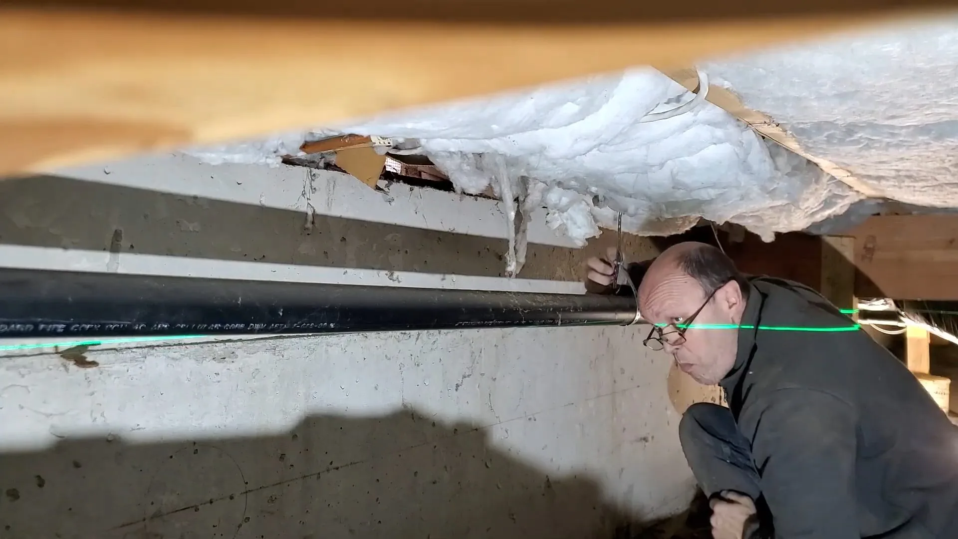 Man in a gray sweater crouched beside a long black drain pipe in a crawl space, with insulation hanging above.