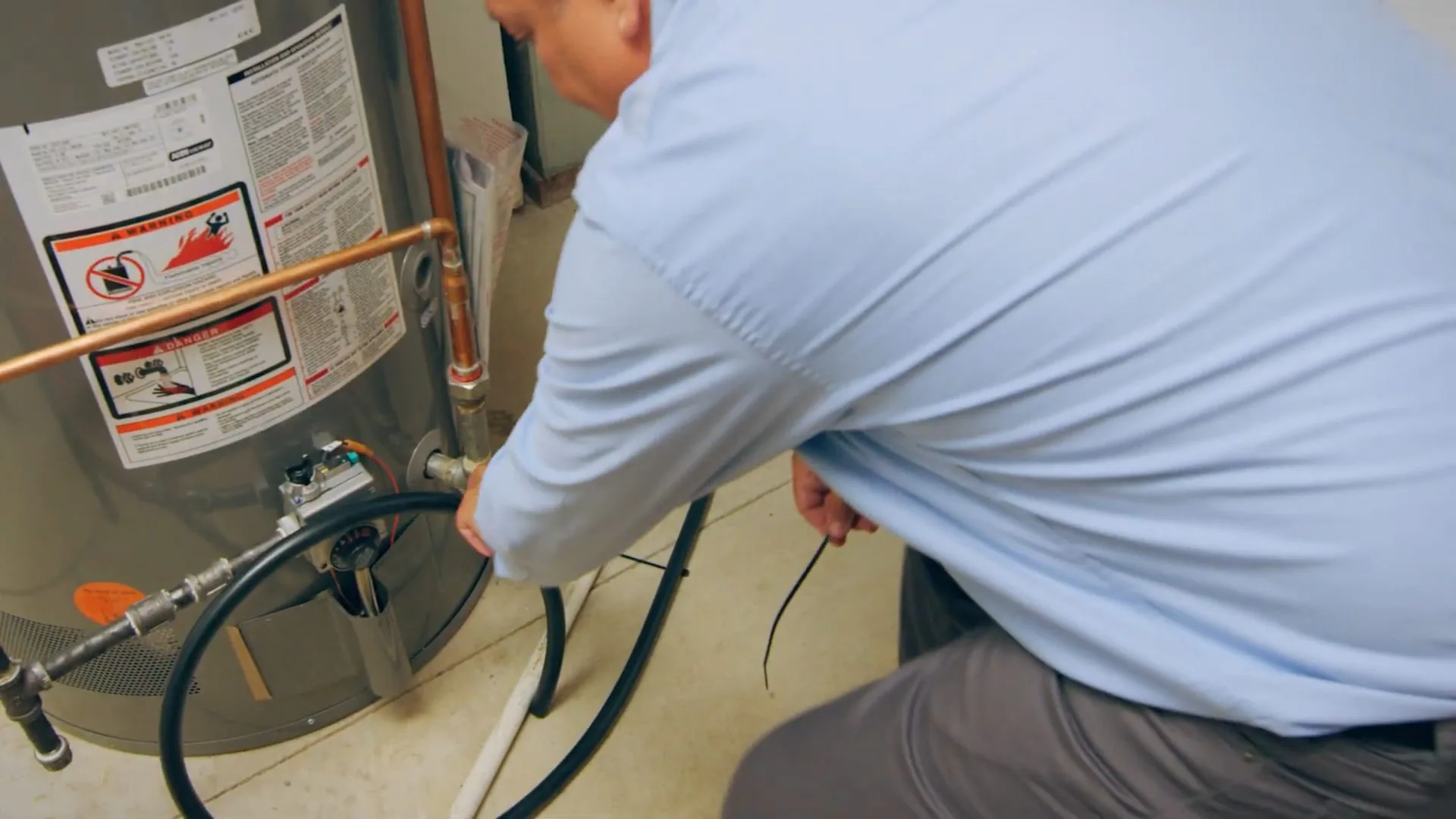 Man in a light blue shirt crouching beside a gas water heater and working on a hose and fitting at its base.