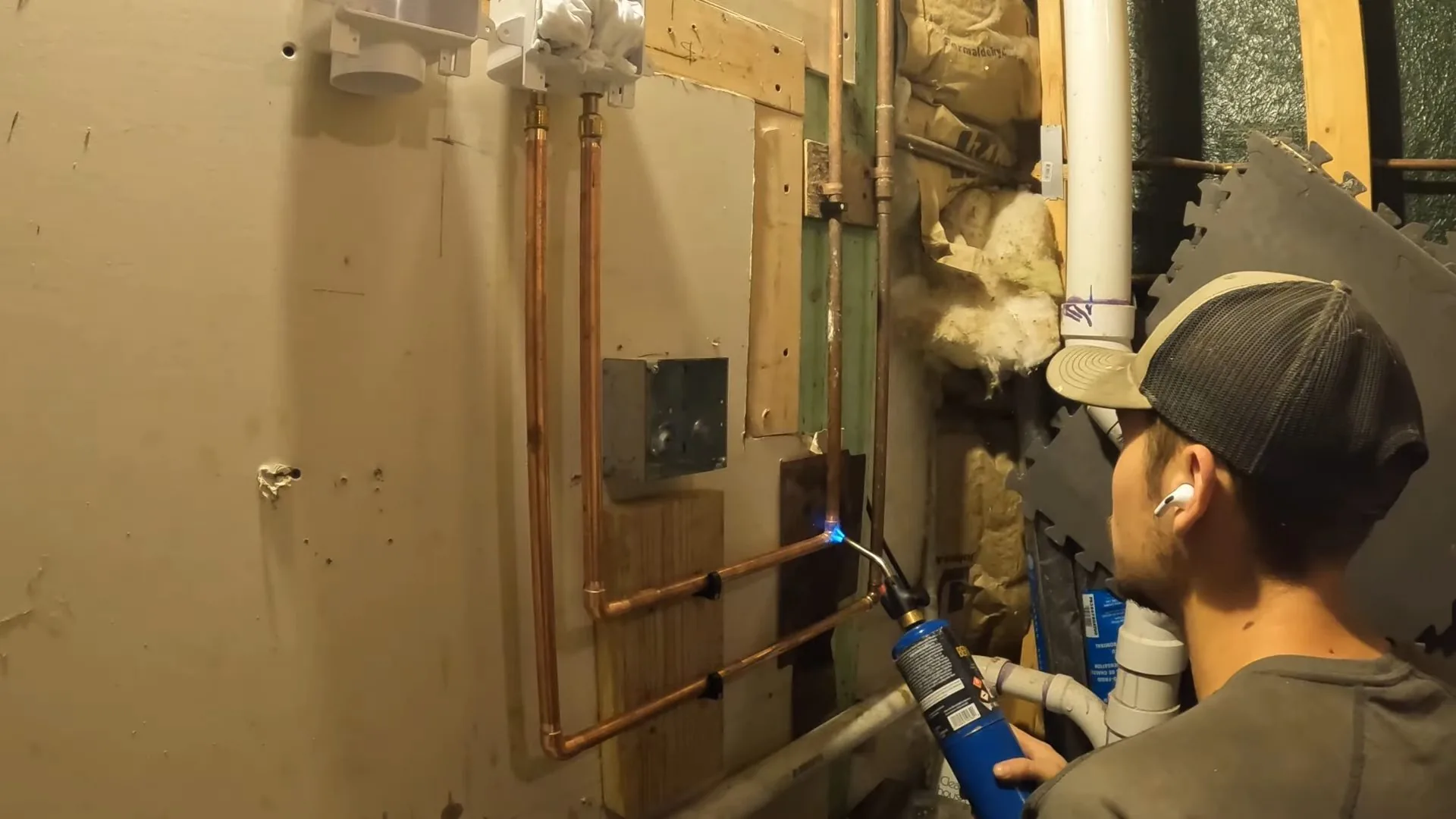 Man in a cap using a torch on copper pipes against a basement wall.