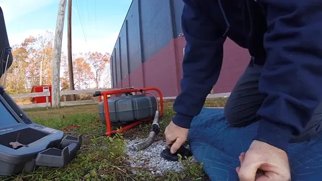Man kneeling on grass beside a gravel-filled cleanout with a red cable reel and a camera case next to him.