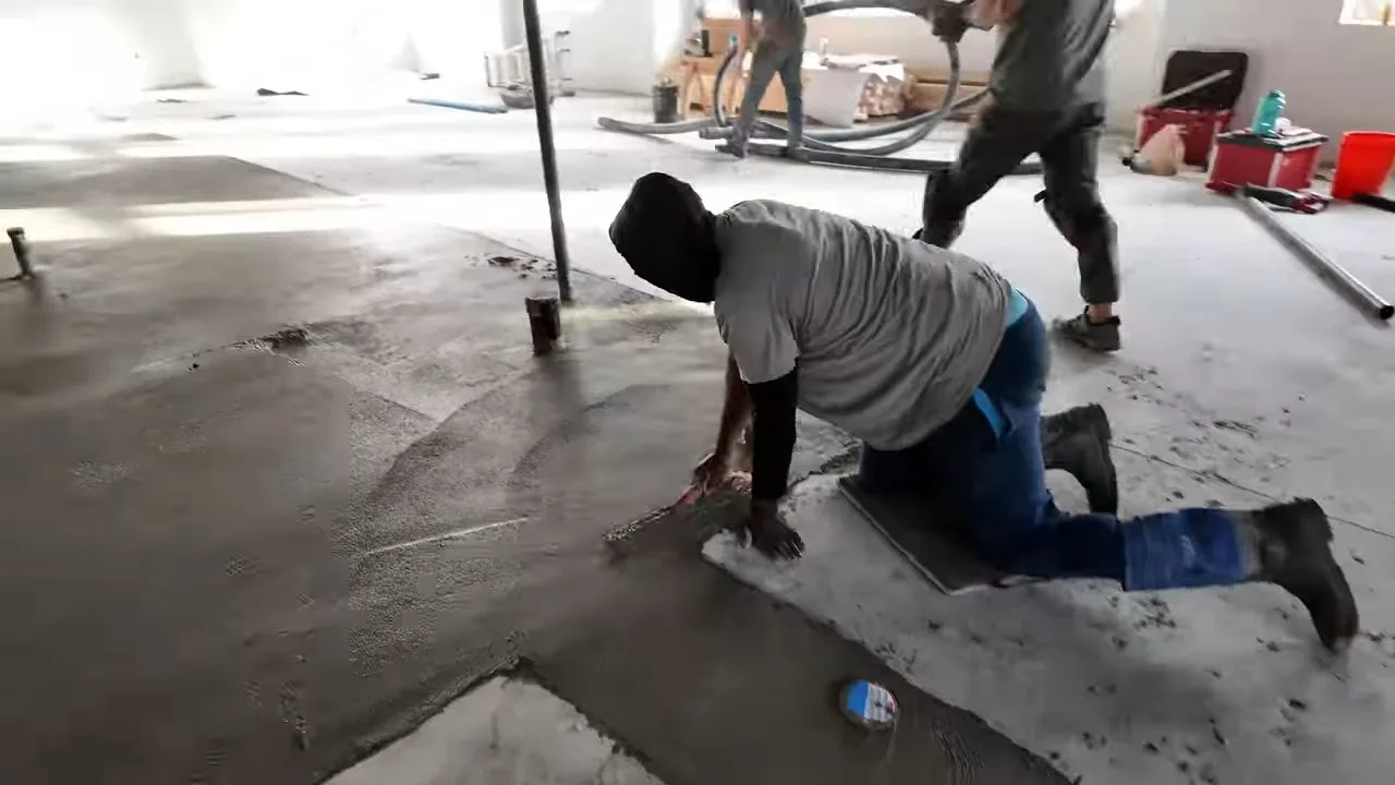 Man on his knees on a gray concrete slab in an open room, checking the surface while another worker stands behind him.