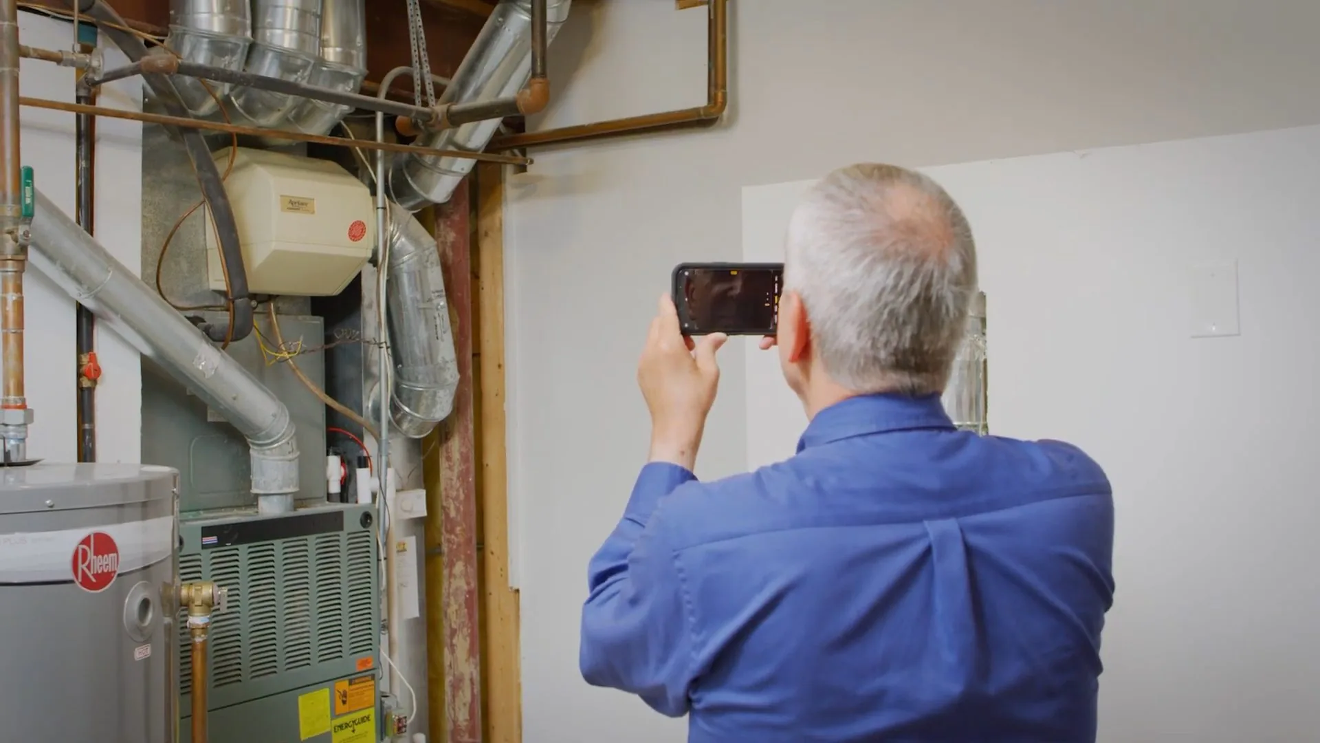 Man in a blue shirt taking a phone photo beside an existing water heater in a utility room.
