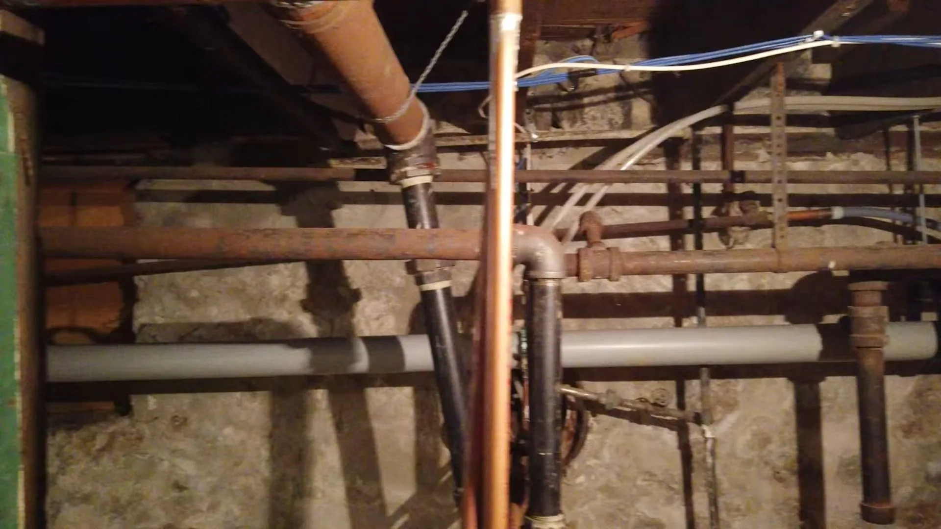 Old copper, iron and plastic pipes running between floor joists in a basement ceiling.