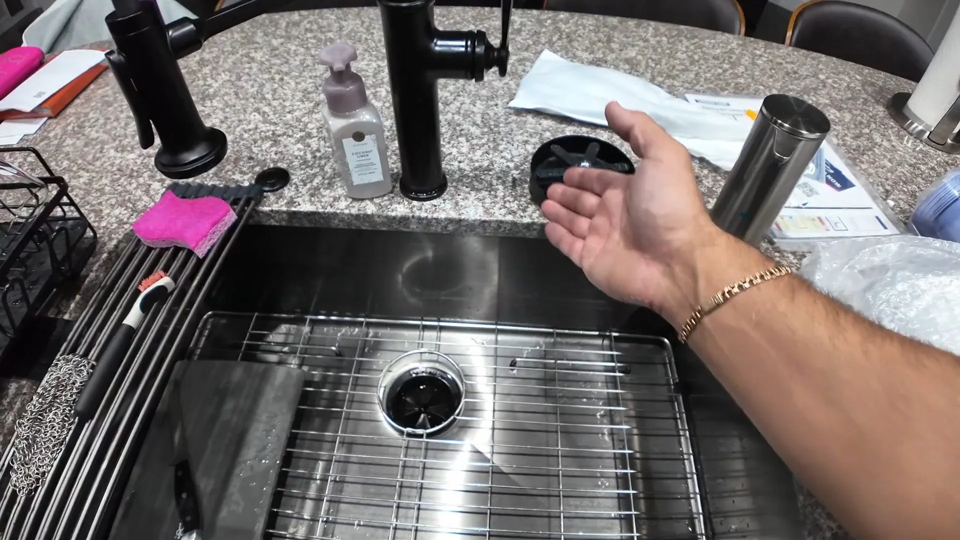 Open hand beside a stainless steel sink whose drain opening leads to a garbage disposal, with a black splash guard and a metal cylinder on the counter.