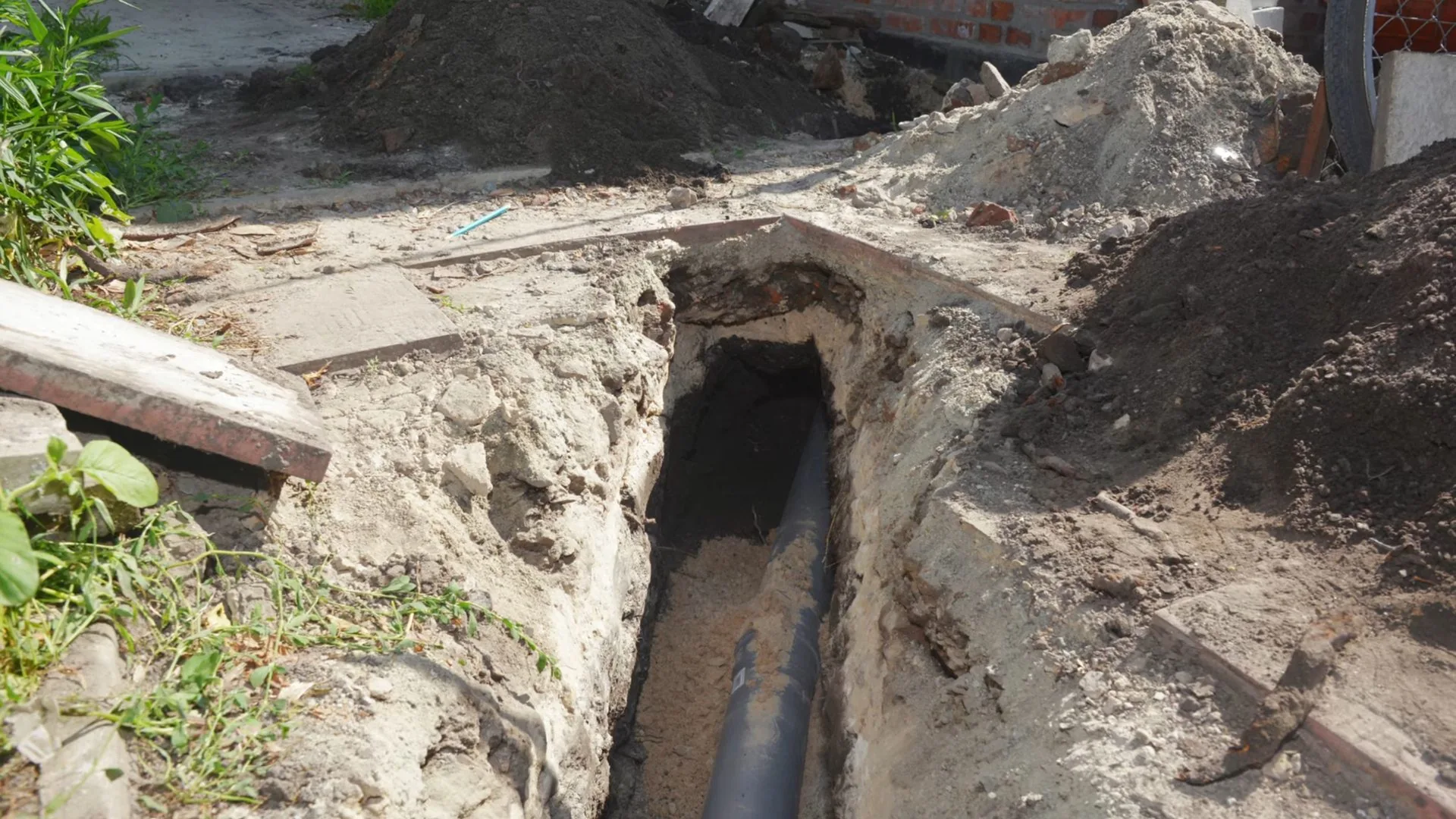 Open trench beside a house wall with a black sewer pipe exposed at the bottom and soil piled on both sides.