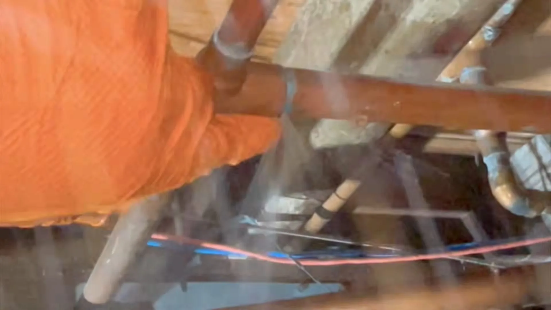 Orange-gloved hand pressed against a copper pipe in an open ceiling while water mists around the fitting.