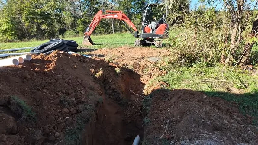 Orange mini-excavator behind an open trench with piled red soil and lengths of white and black pipe beside it.