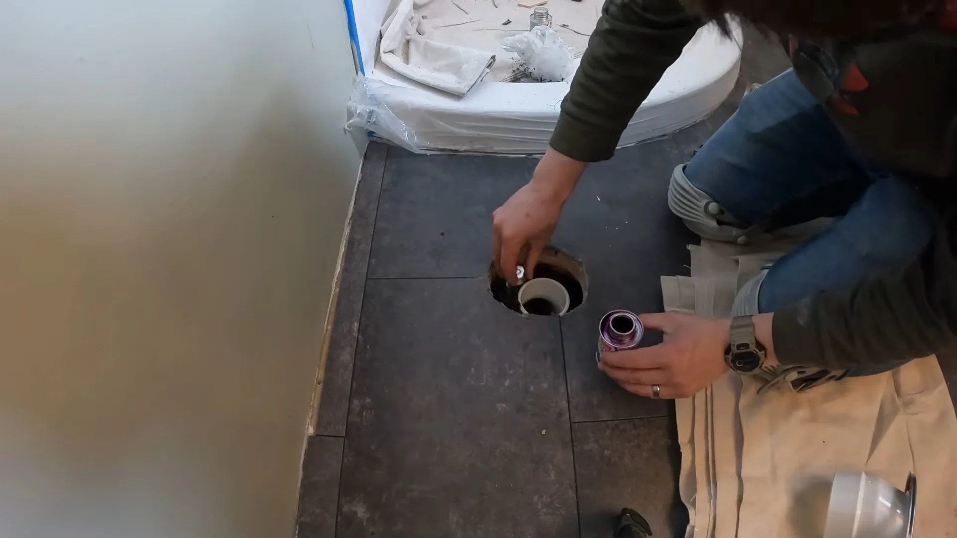 Person kneeling on a tile floor beside an open floor drain flange, with a white bowl at the top of the frame.