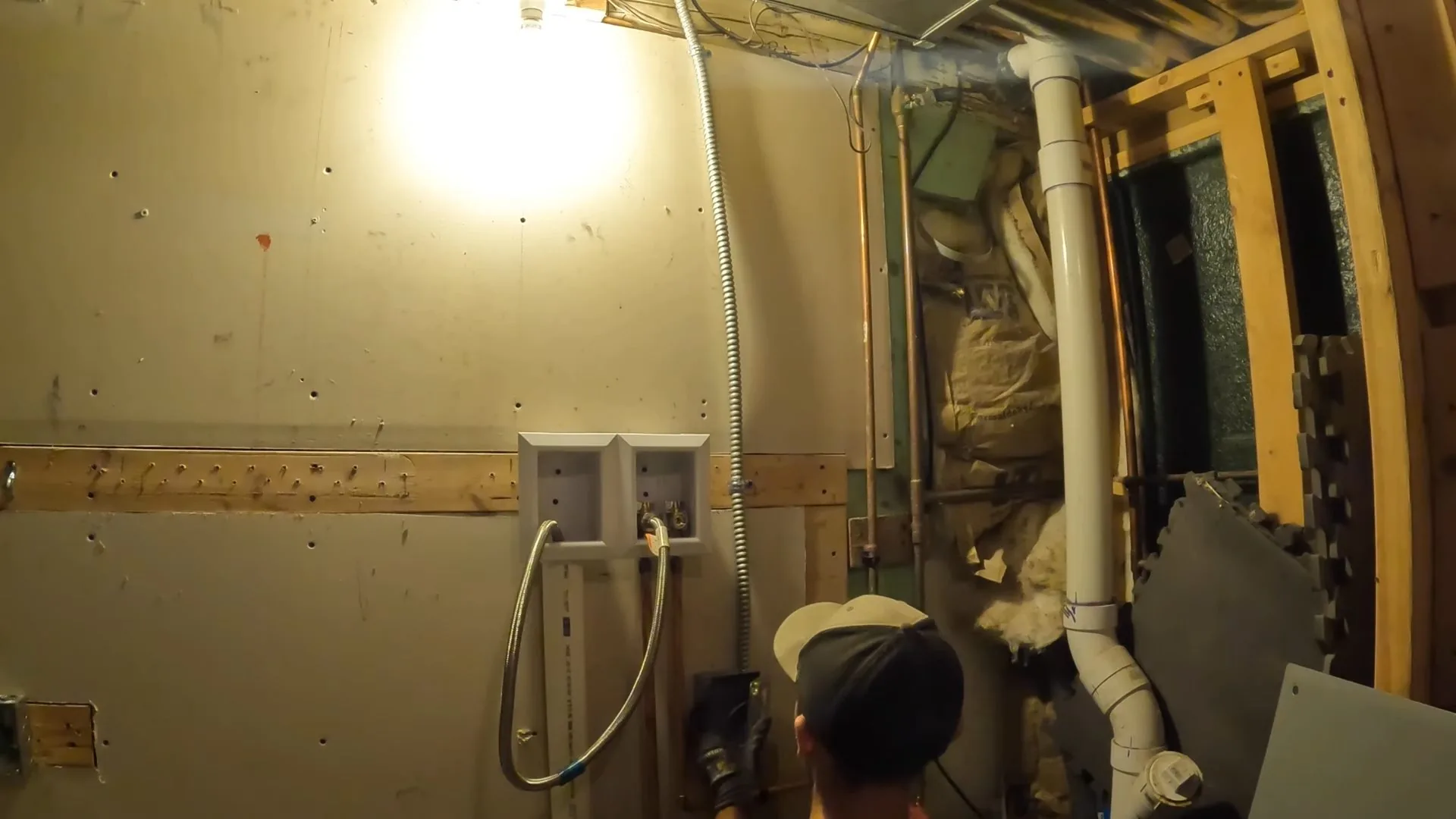 Person in a cap working beside a white drain stack in a framed basement wall.