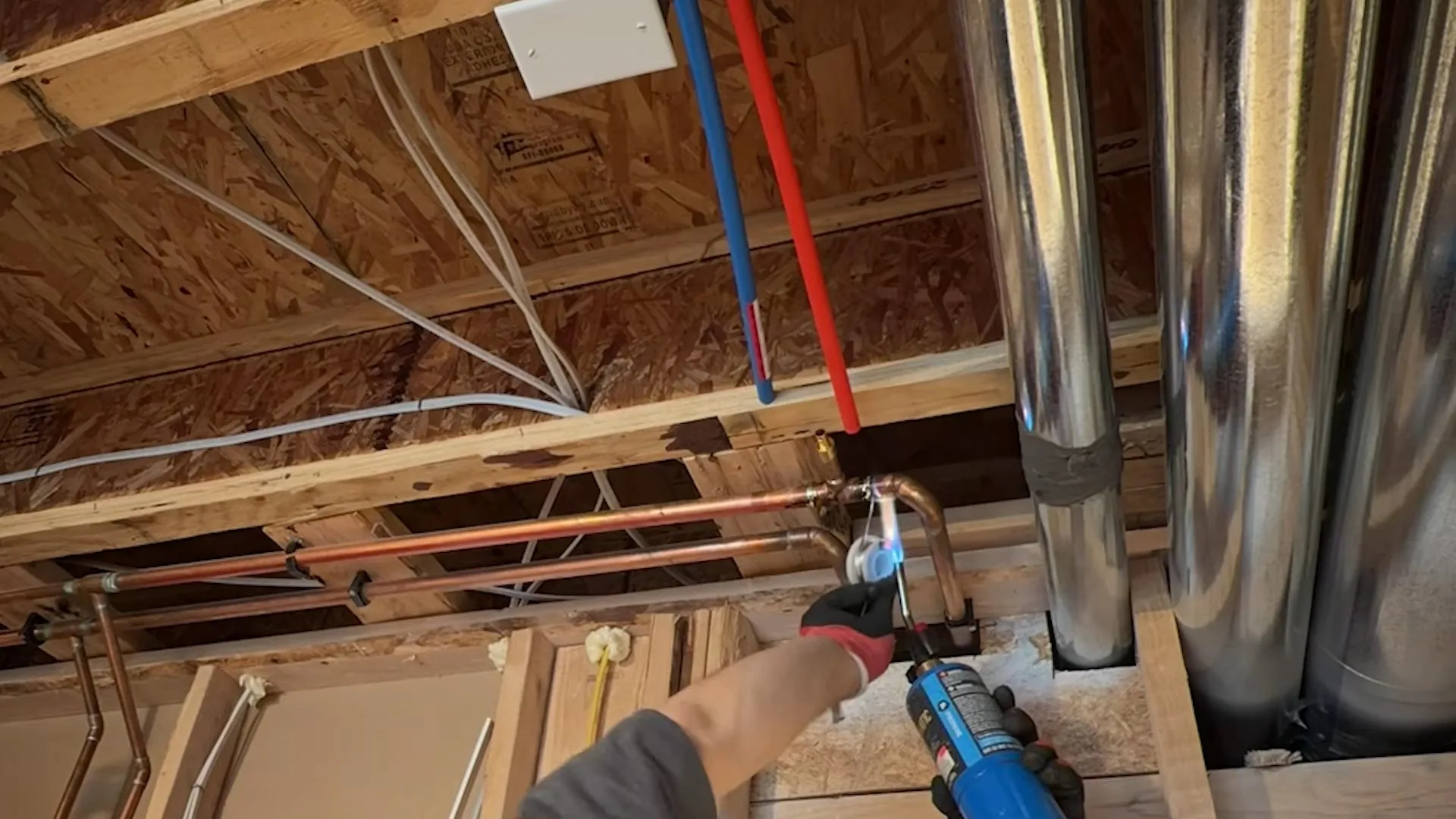Open basement ceiling joists with red and blue PEX lines and a hand holding a crimp tool at a copper connection.