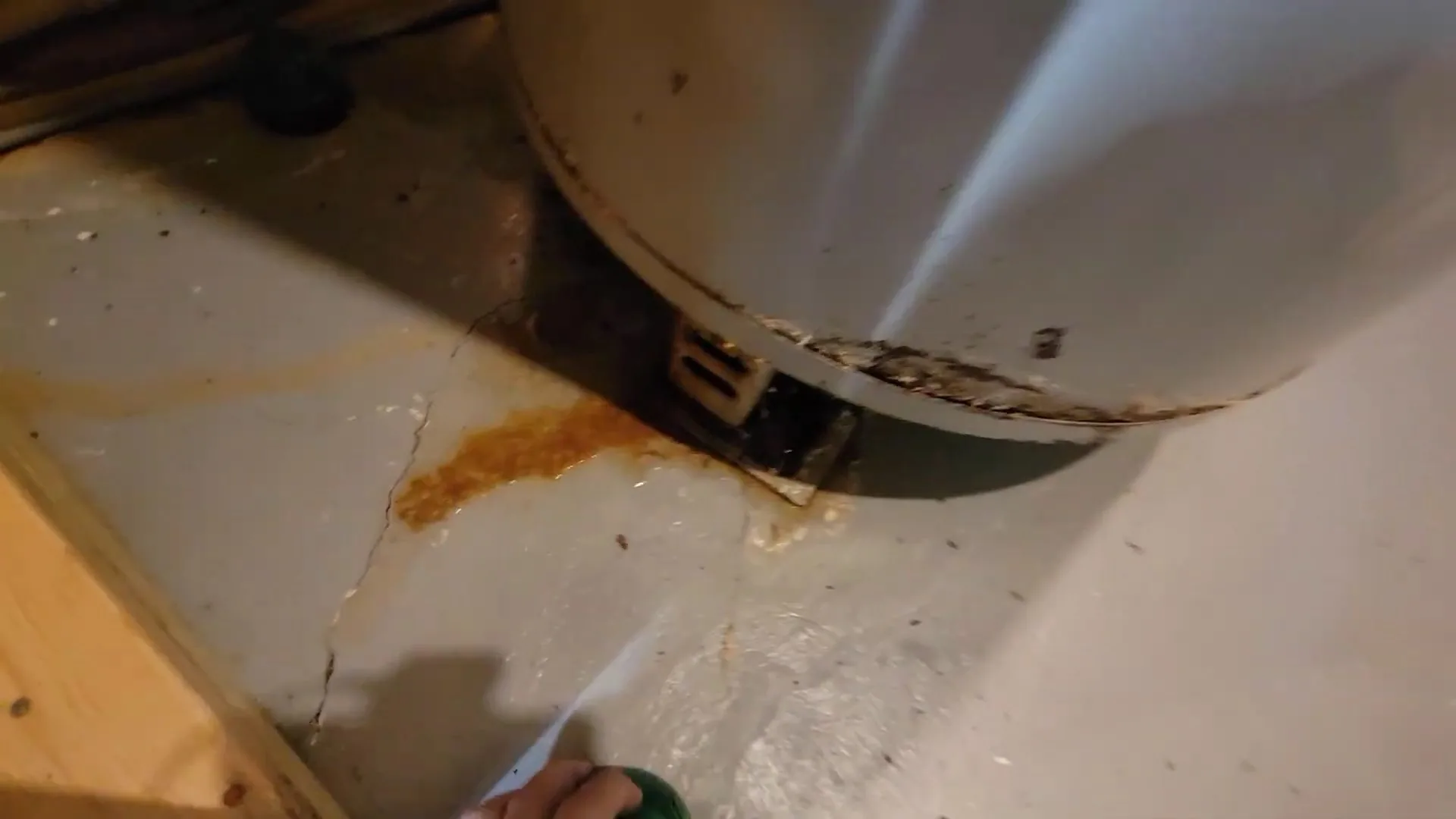 Bottom of a white tank water heater with rust staining on the floor and a jet of water coming from its base.