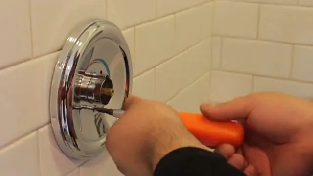 Hand with an orange screwdriver removing the chrome trim plate from a shower valve against white tile.