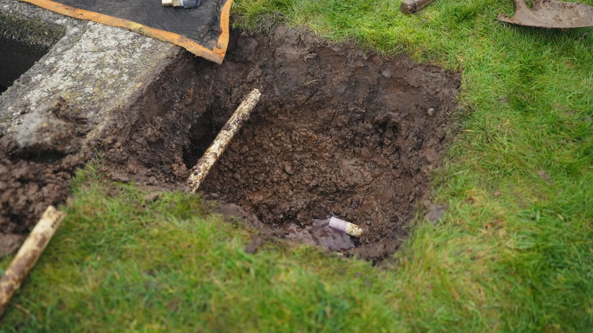 Square excavation in a grassy yard with a shovel handle resting in the hole.
