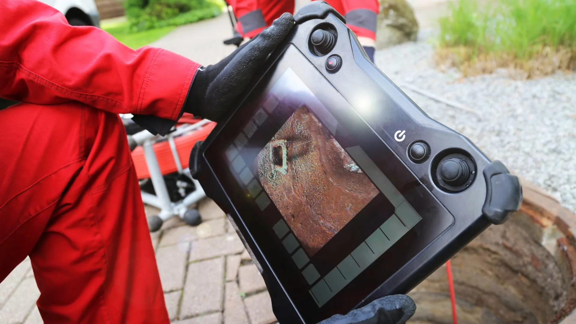 Technician in red coveralls holding a handheld screen that shows the inside of a pipe.