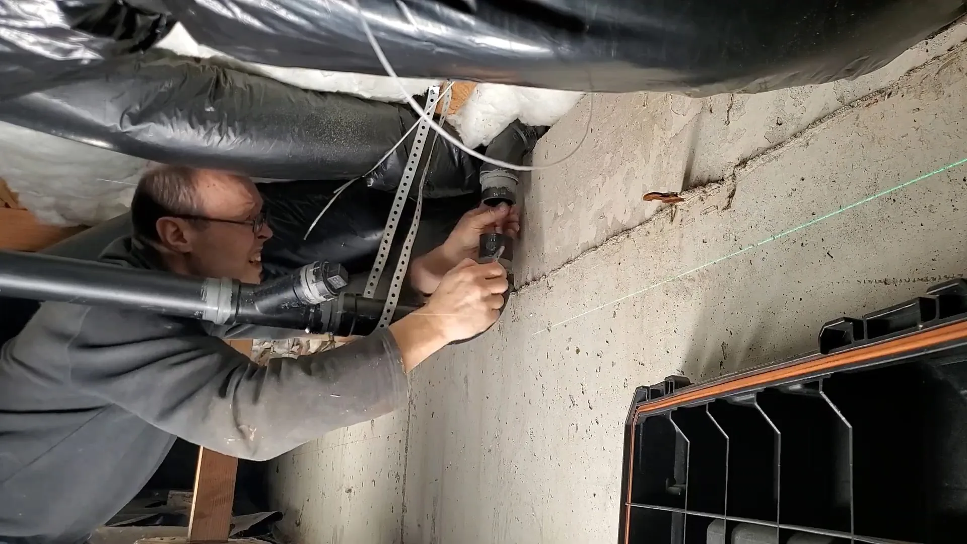 Man with glasses reaching up to a pipe in a low crawl space with ductwork above him.