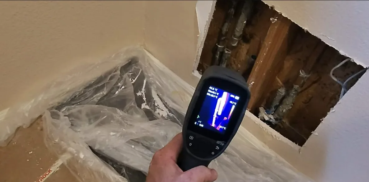 Hand holding a thermal imaging detector in front of an opened wall and a plastic-covered floor corner.