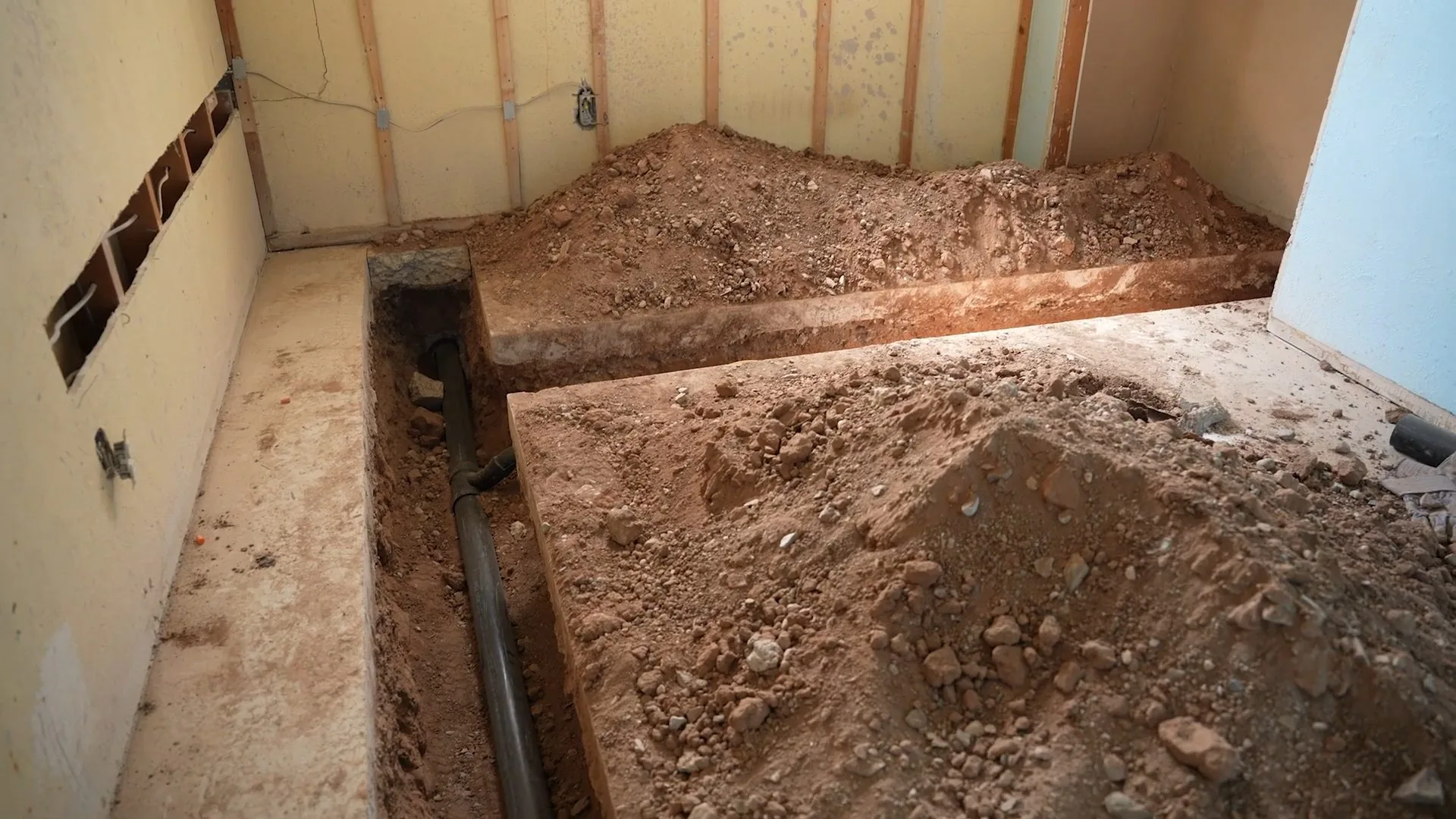 Dirt-filled trench cut into the floor of a room with bare wall studs, with a pipe visible in the trench.