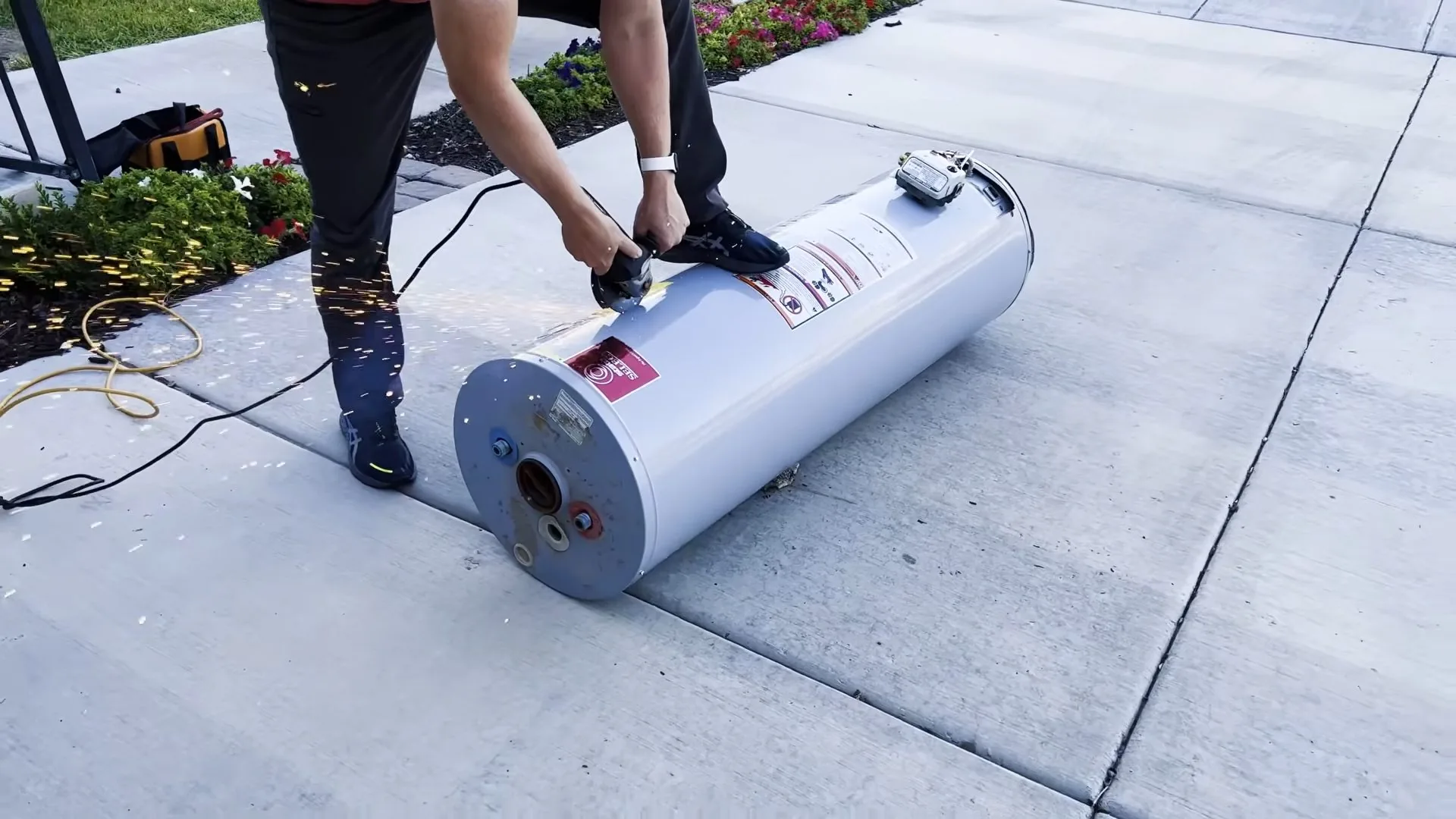 Two people standing over a white water heater tank lying on its side on a concrete patio.