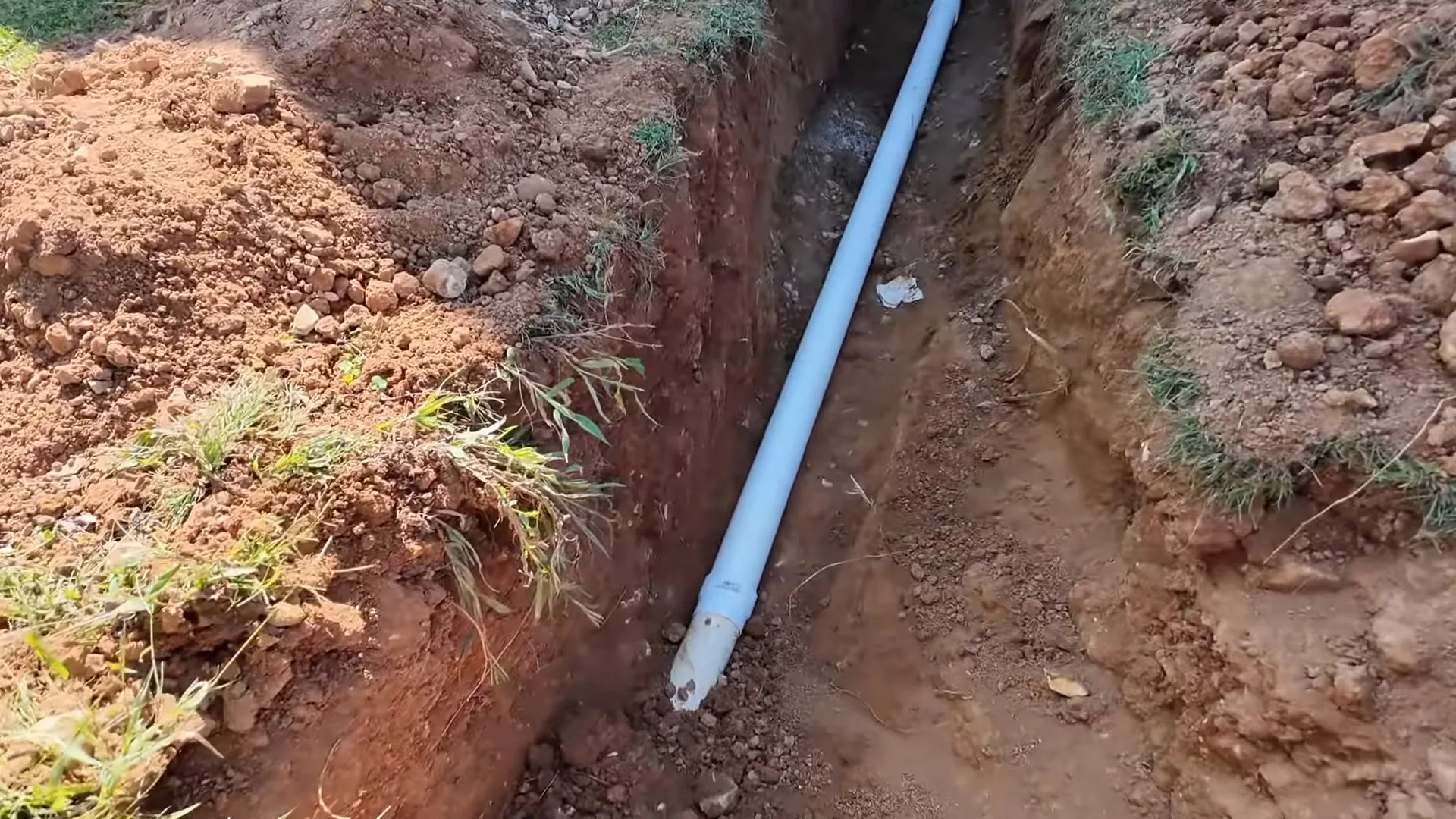 White PVC pipe laid in a narrow dirt trench with grass along its edges.